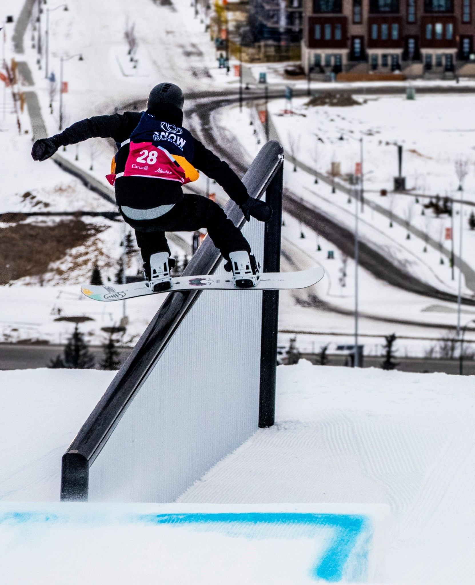 Snowboarder sliding a rail feature on a snowy slope with urban backdrop and winding road.
