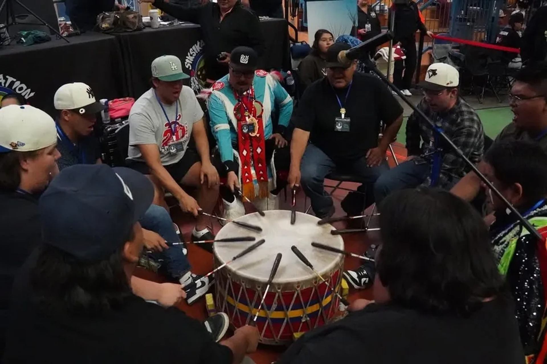 Drum circle musicians sit around large powwow drum, striking in unison indoors.
