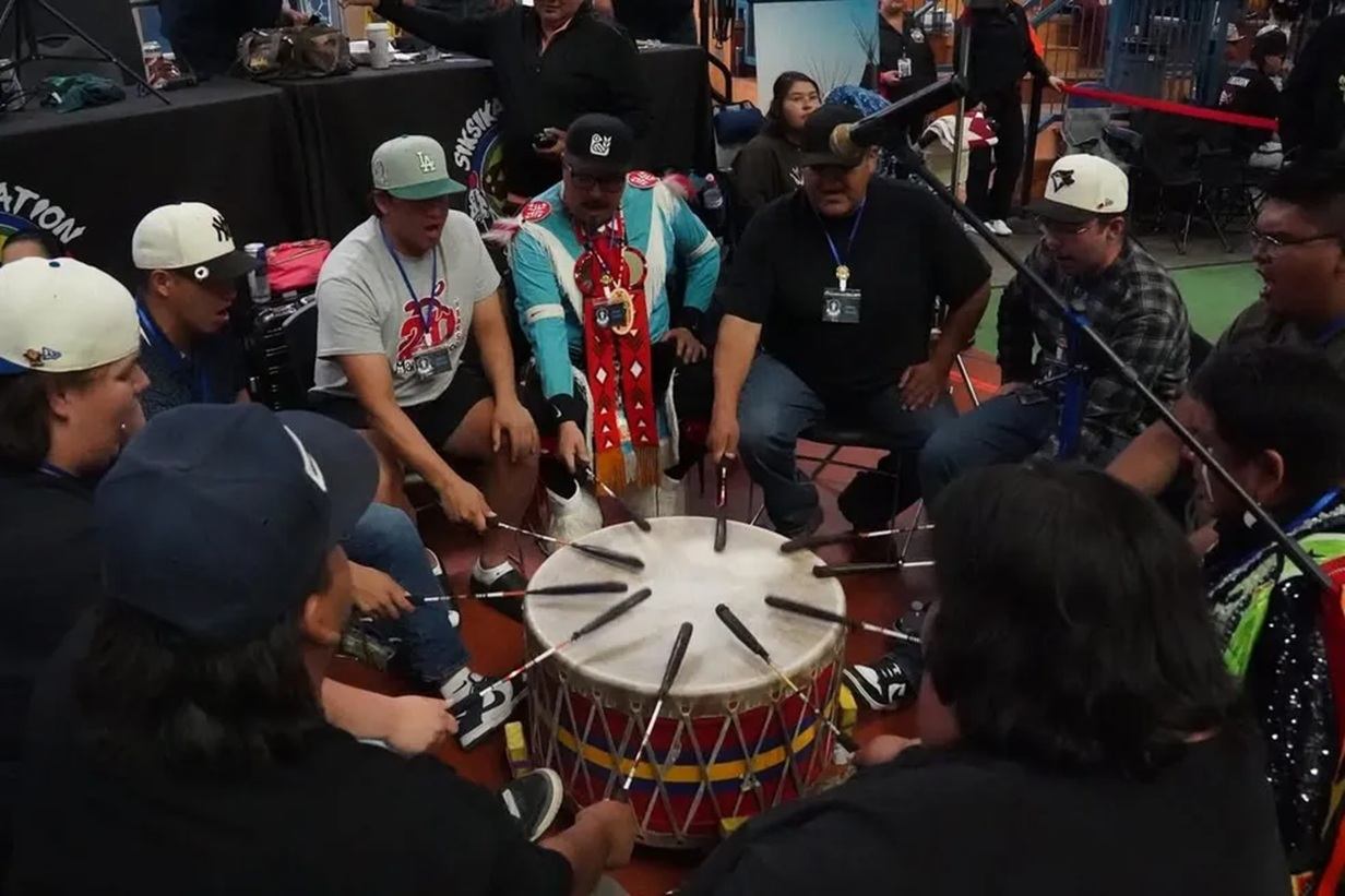Drum circle musicians sit around large powwow drum, striking in unison indoors.