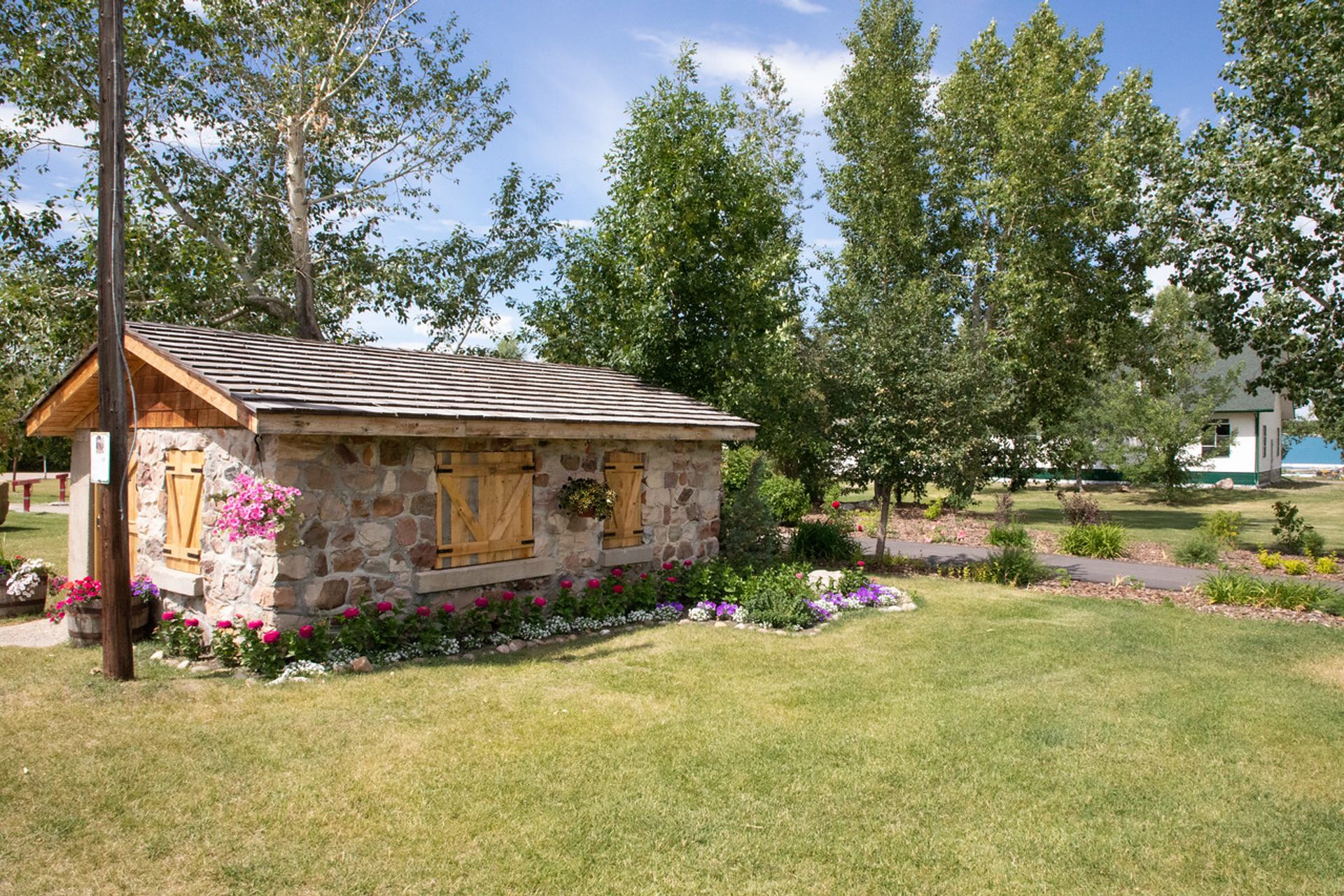 Stone garden shed with wooden shutters surrounded by flowers, trees and mowed lawn.