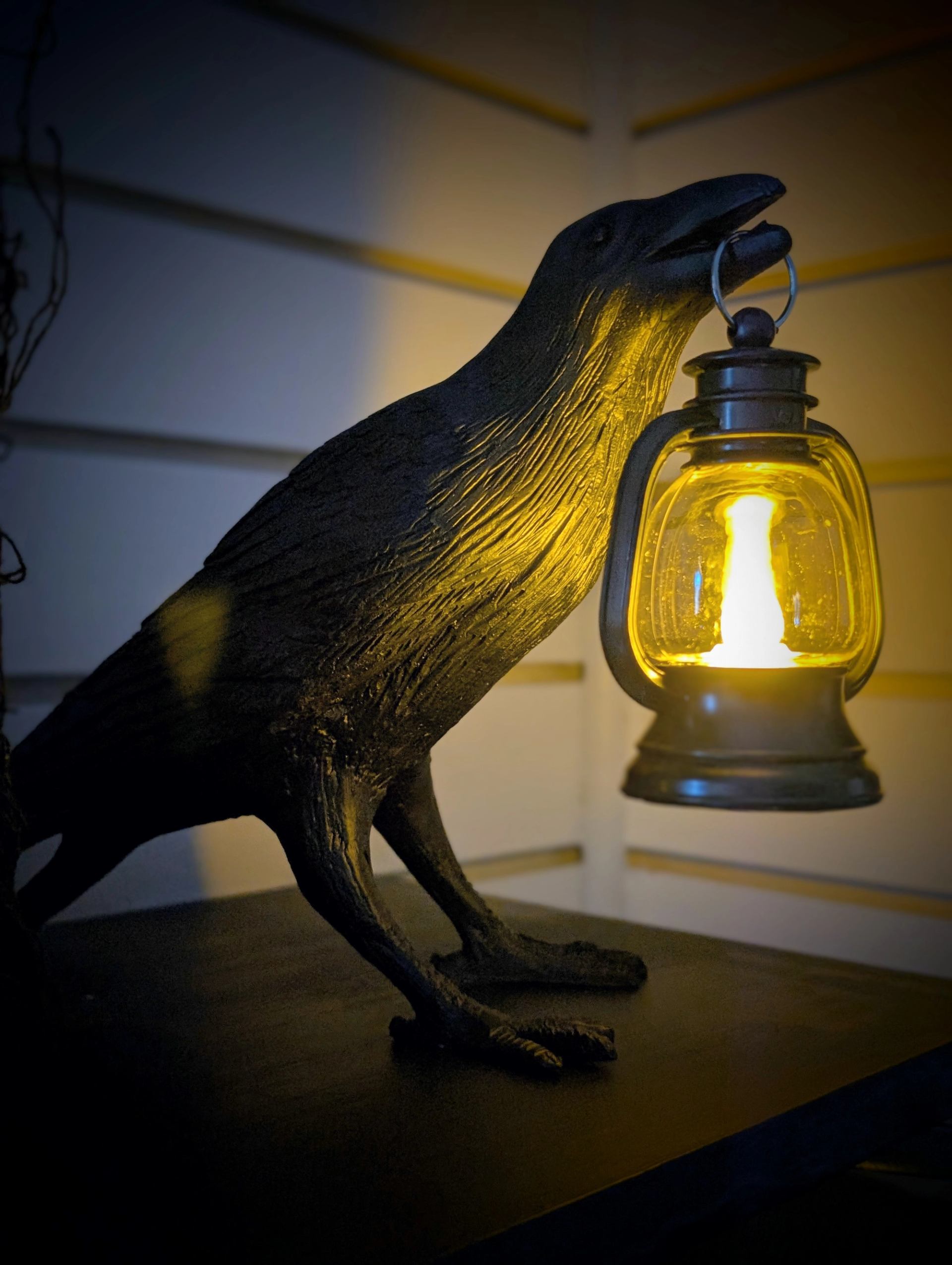 Black raven sculpture holding a glowing lantern against a wooden wall backdrop.
