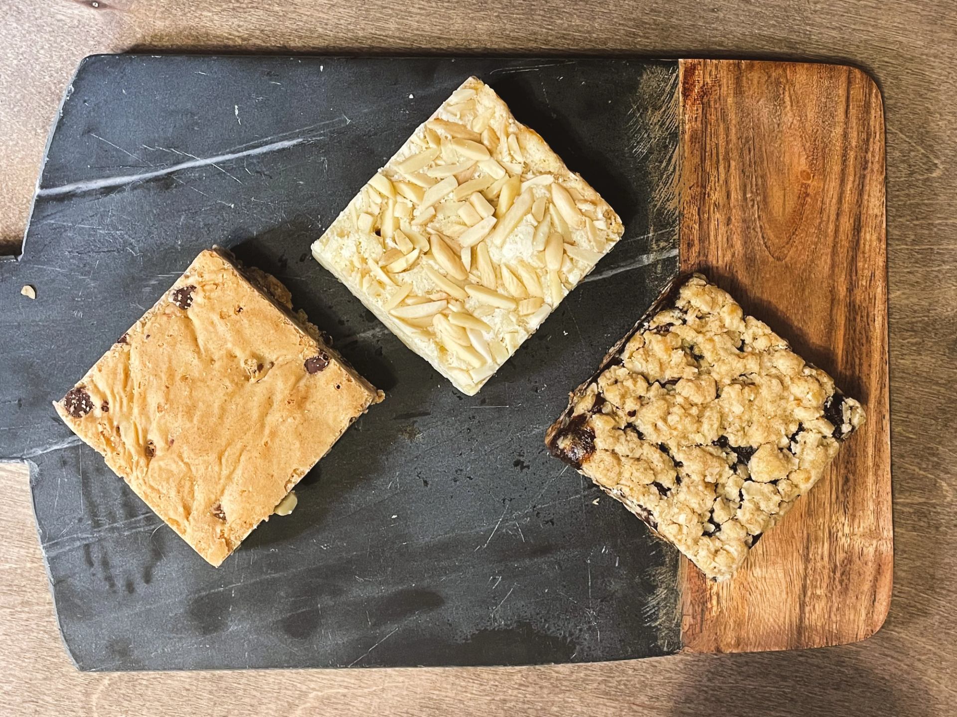 Three assorted square dessert bars on a black and wood serving board.