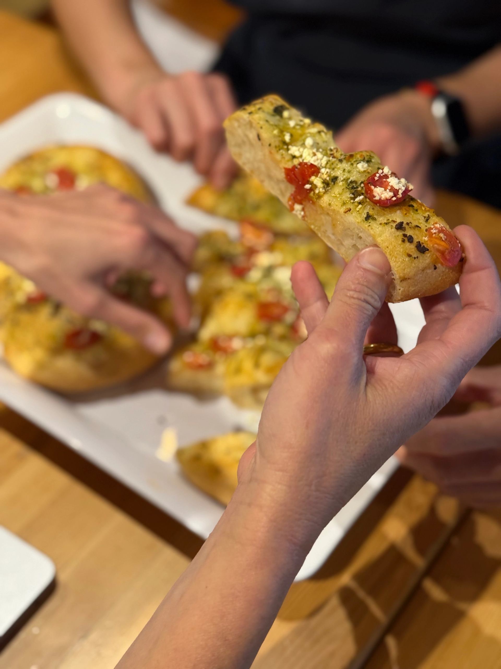 Hands reaching for garlic bread topped with tomatoes and herbs on a white plate.