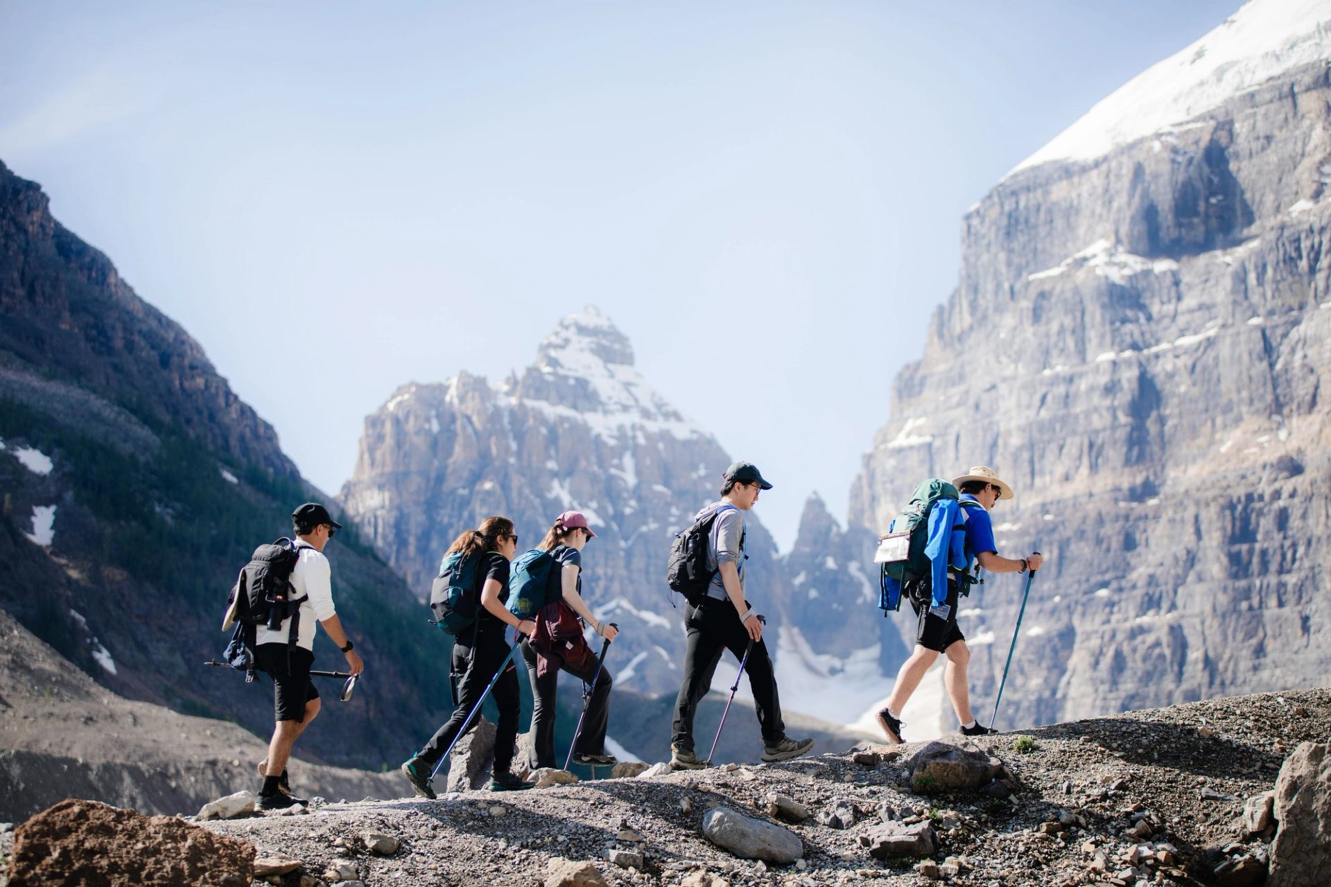 Hikers with backpacks and poles walk along rocky trail below snow-capped mountain peaks.
