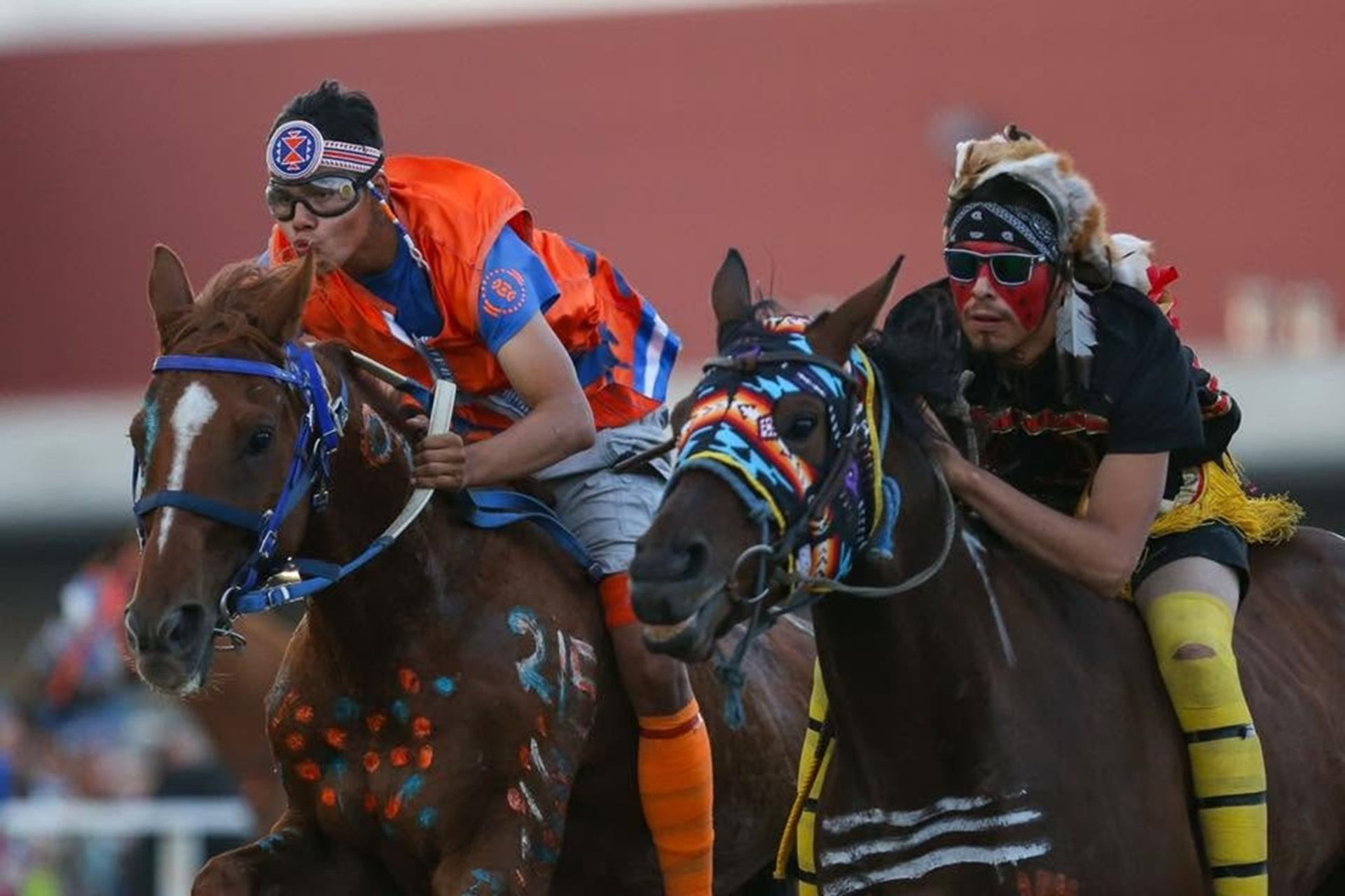 Bareback riders in colourful regalia race decorated horses on a track.