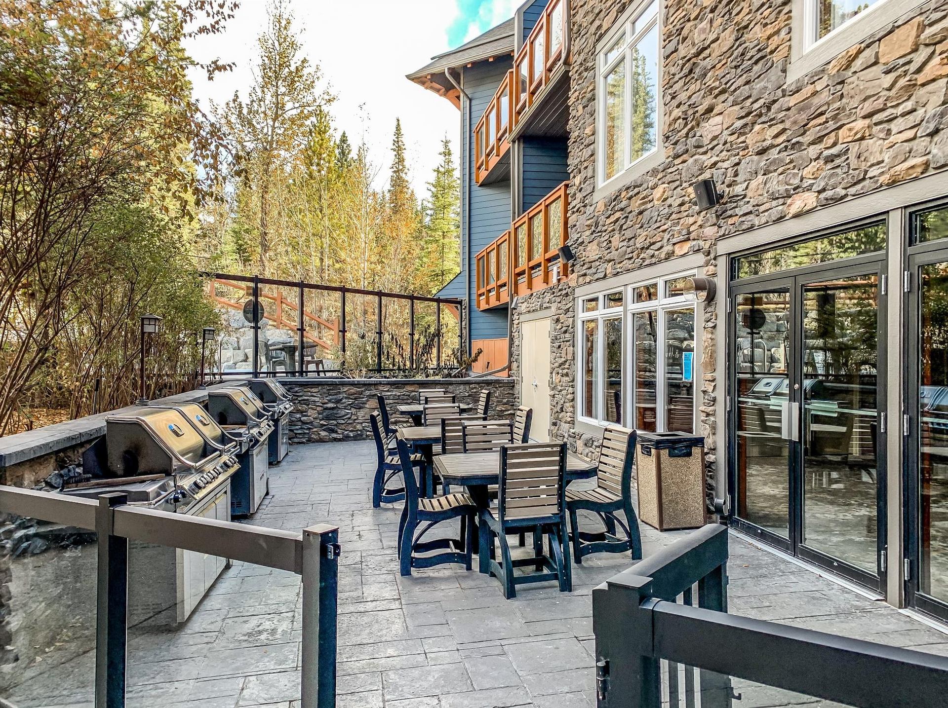 Outdoor patio with barbecue grills, wooden tables and chairs beside stone building and trees.