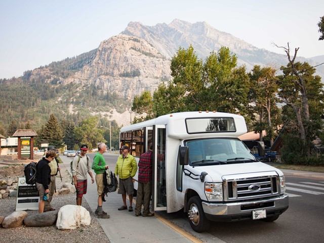 Day hikers board a small shuttle bus in Waterton, with rocky mountains rising behind.