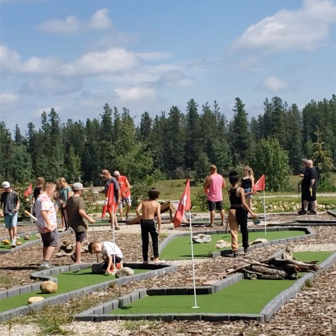 Guests playing mini-golf at Pinnacle Trails Resort under a sunny sky with forest backdrop.