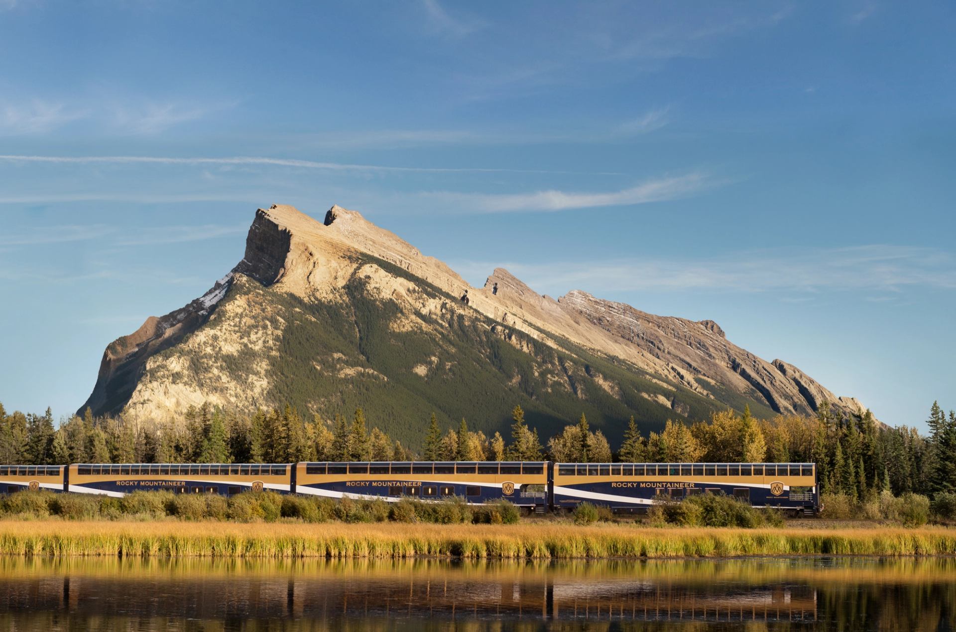 Rocky Mountaineer train traveling beside a calm river with grassy banks and a mountain backdrop.