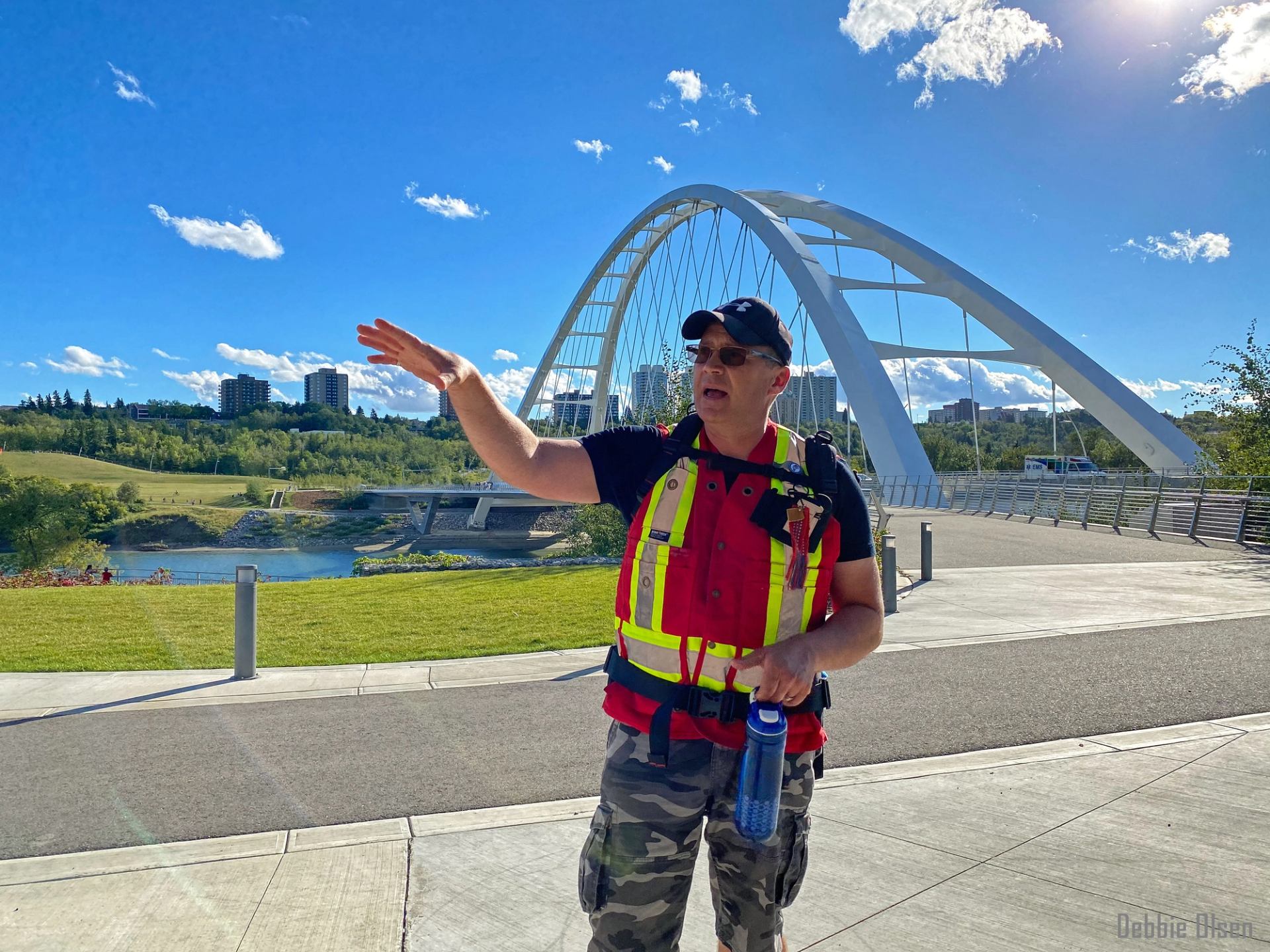 A Métis guide is talking about the local fur trade history near a white arch bridge on a sunny day.