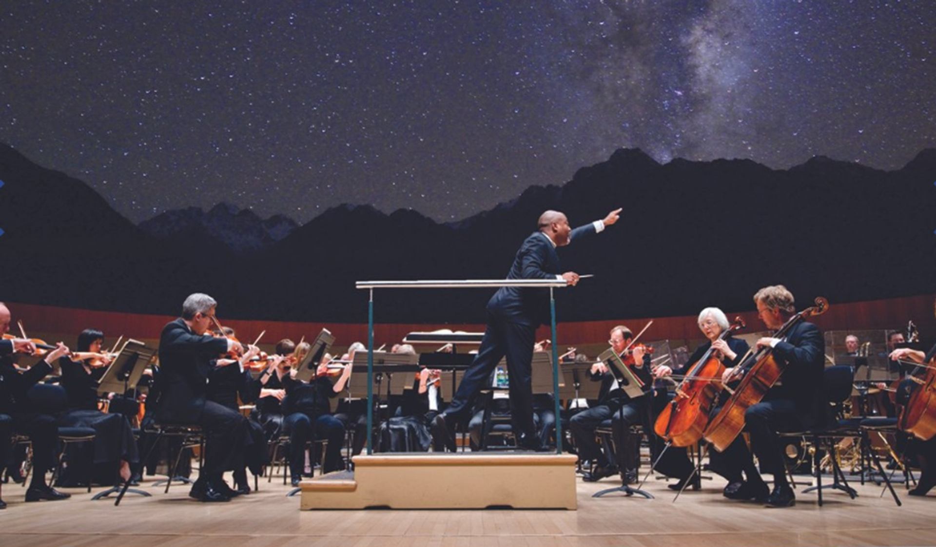 Symphony orchestra performs onstage as conductor leads, with starry night backdrop projection.