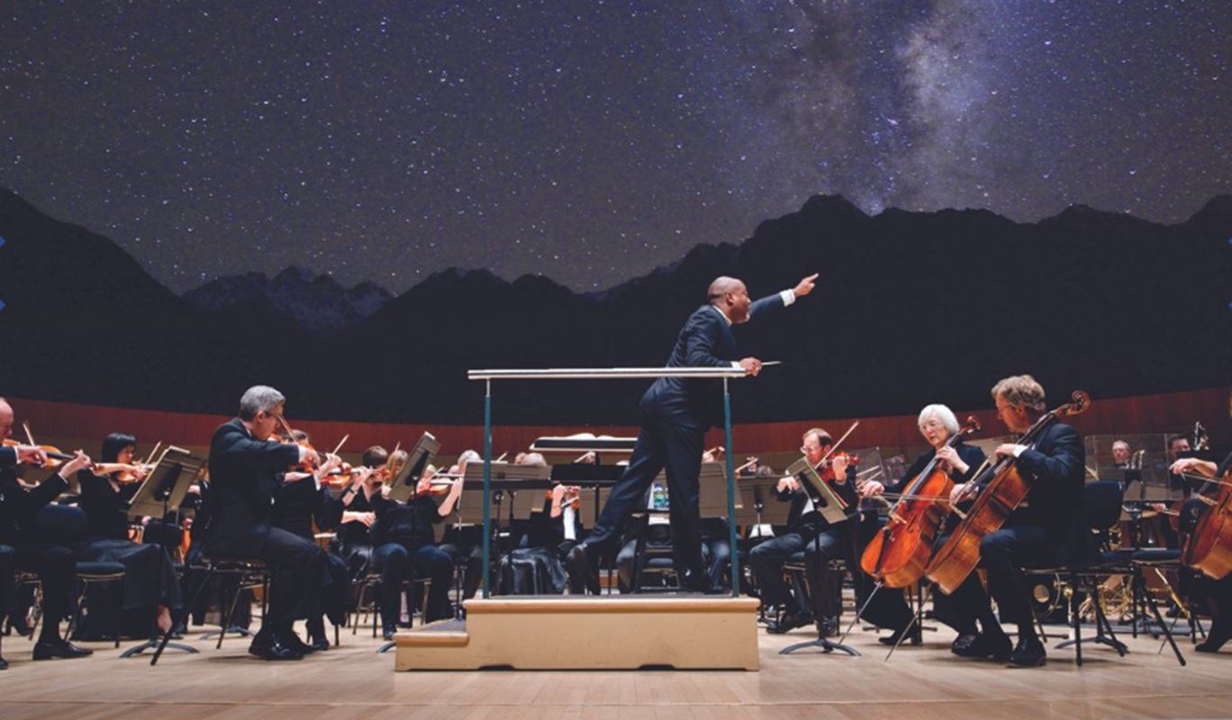 Symphony orchestra performs onstage as conductor leads, with starry night backdrop projection.