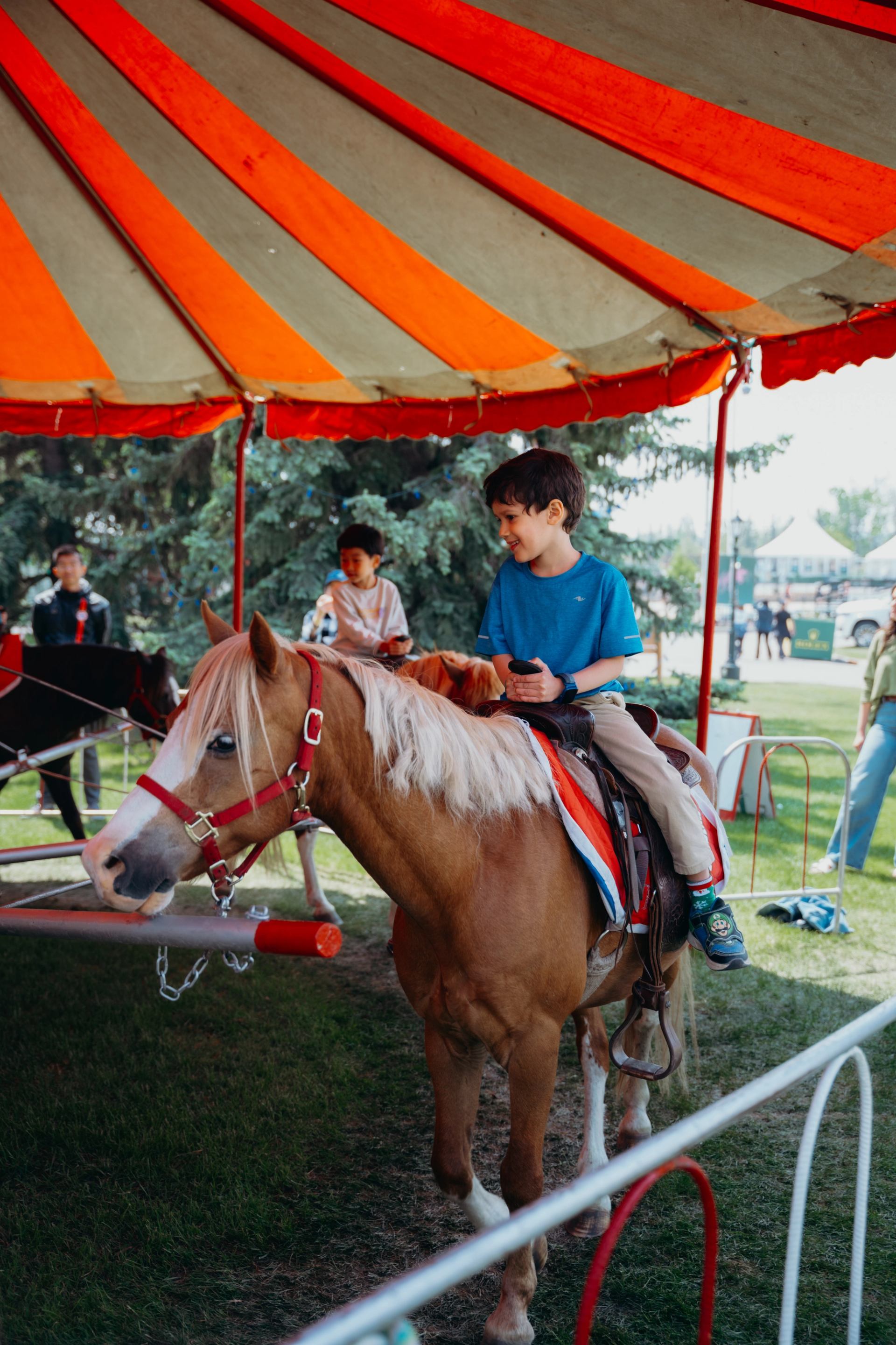 A boy smiles while riding a pony under a striped tent at a fair.