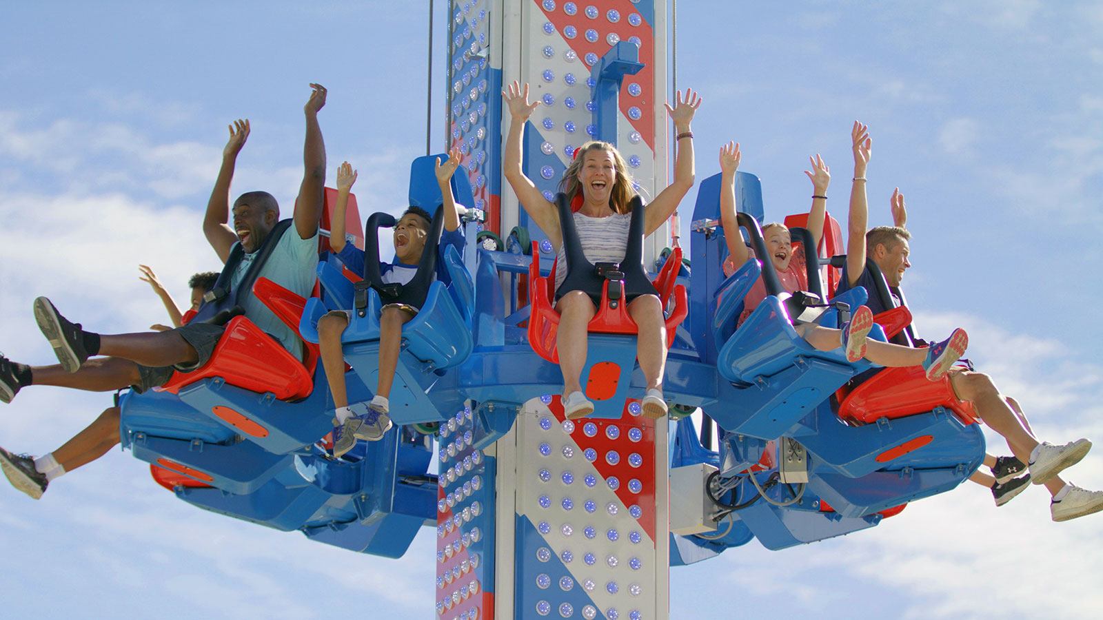 People ride a colourful drop tower amusement ride, seats raised high against blue sky.