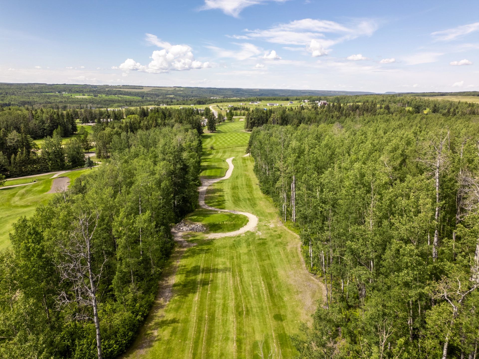 Aerial view of a golf course fairway with a winding cart path, bordered by dense green forests, under a blue sky.