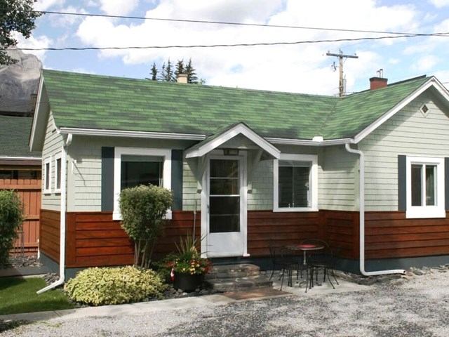 A small, cozy house with green roof, light gray siding, and red-brown lower trim. A front garden, black patio table, and clear sky create a welcoming feel.