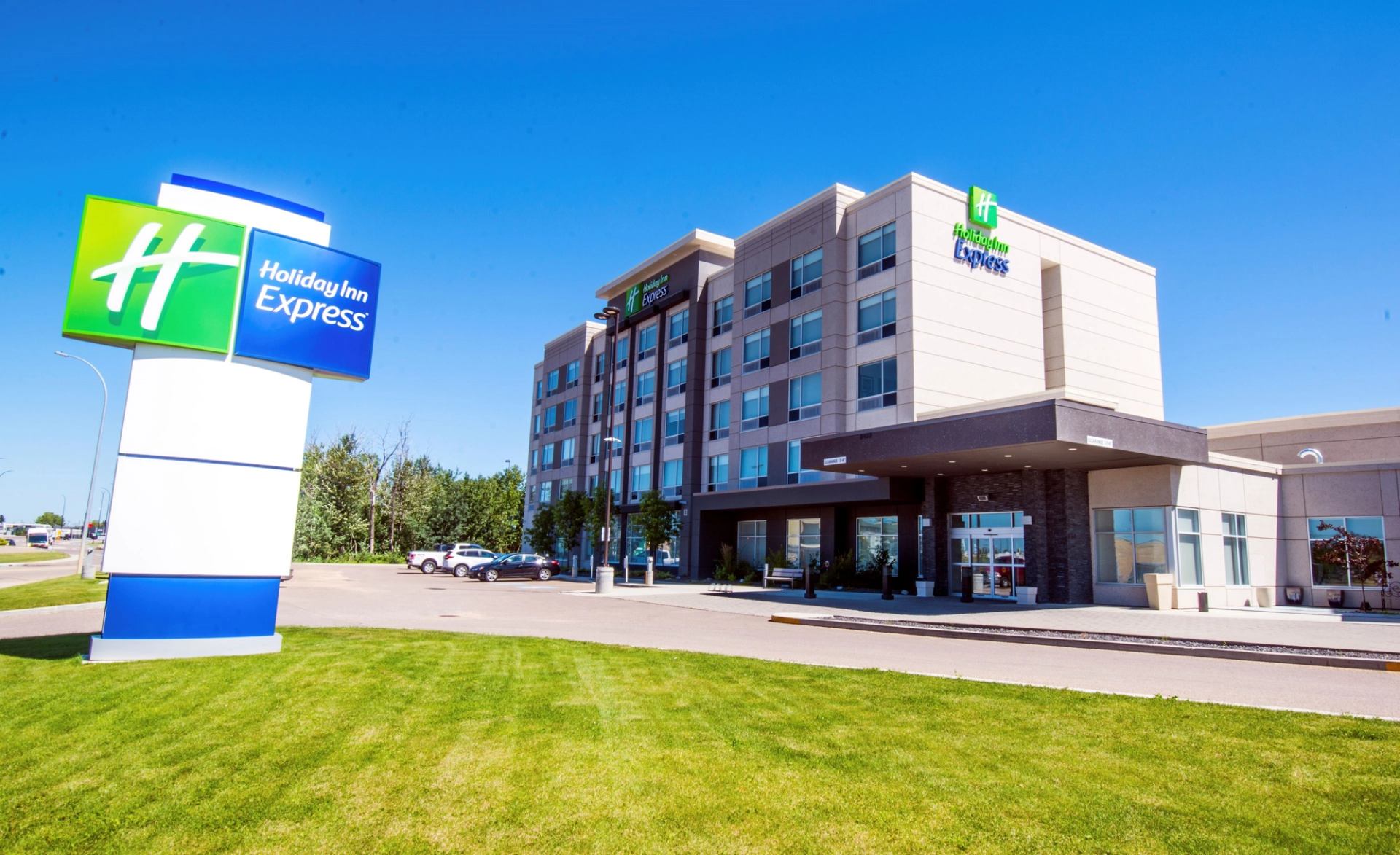 Modern hotel exterior with five stories, featuring sleek lines and large windows under a clear blue sky. The Holiday Inn Express sign is prominent on lush green grass.