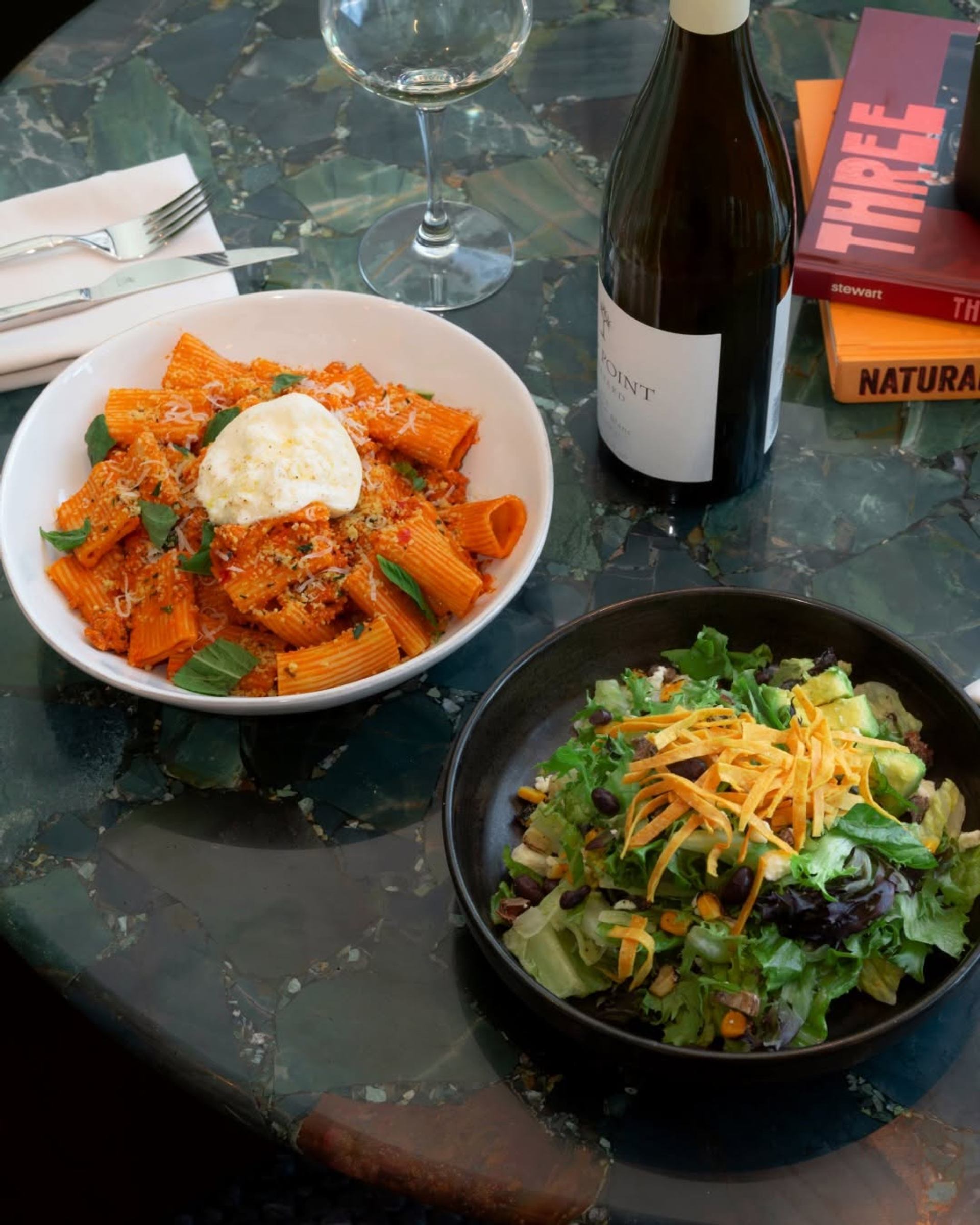 Rigatoni pasta and a fresh green salad are served beside a bottle of wine on a marble table.