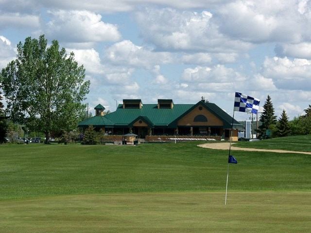 Golf green with flag, clubhouse in background, and trees under a partly cloudy sky.