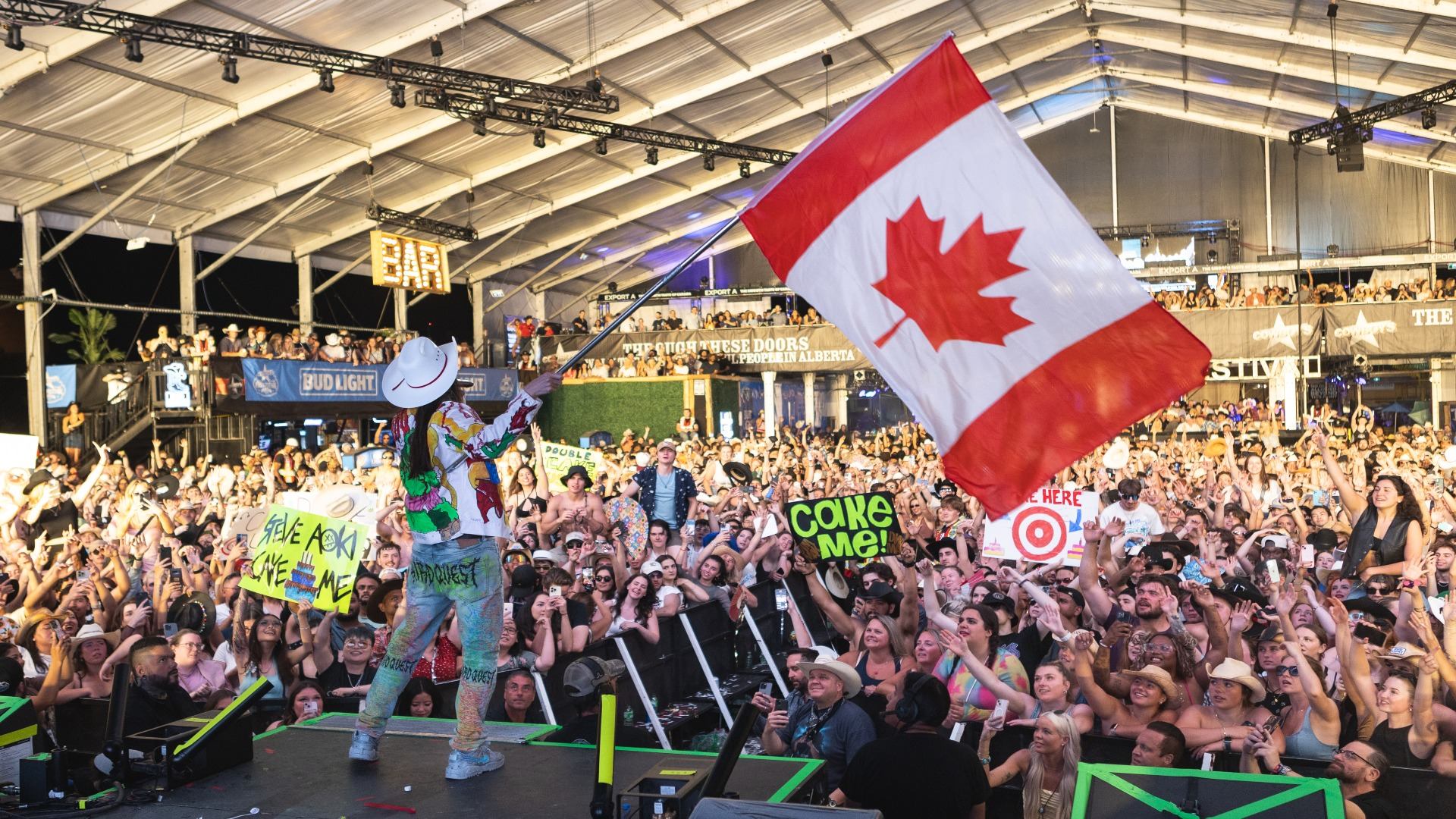 A performer on stage waves a large Canadian flag over a dense, cheering crowd at a concert.