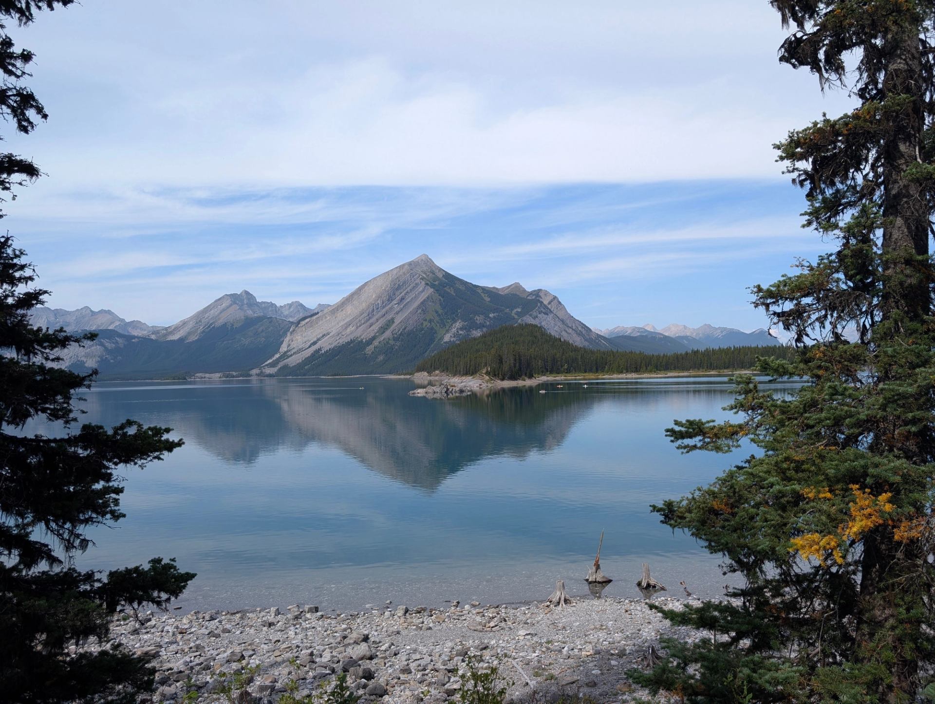 Mountain lake with rocky shore, evergreen trees and peaks reflected in calm water.