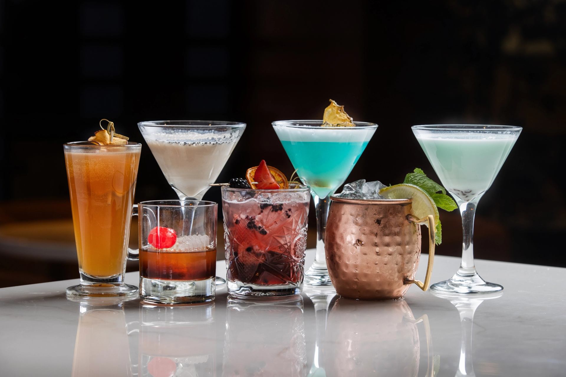 Assorted colourful cocktails in various glasses and a copper mug on a reflective bar.