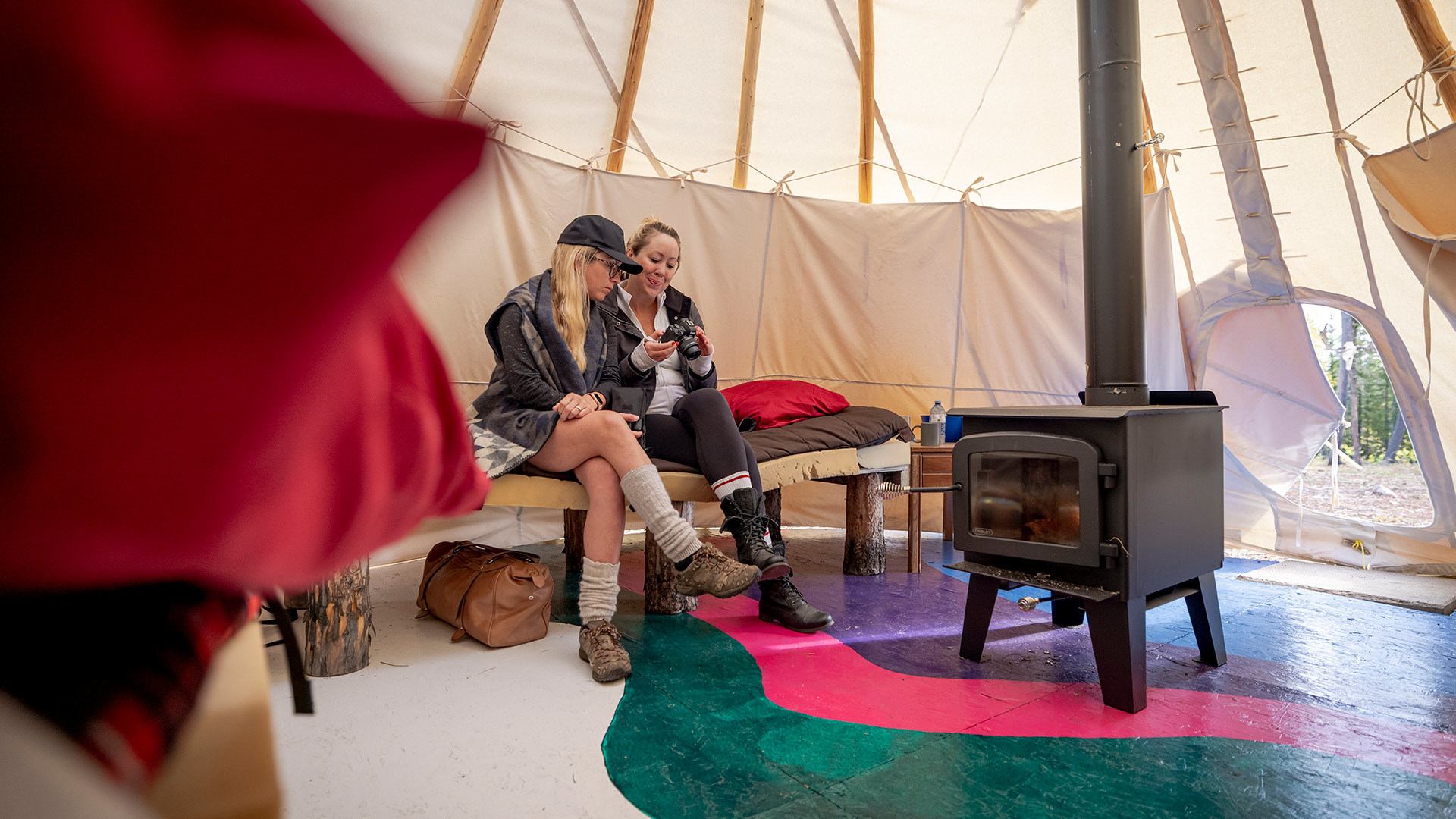 Two women sit on a bench near a wood stove.