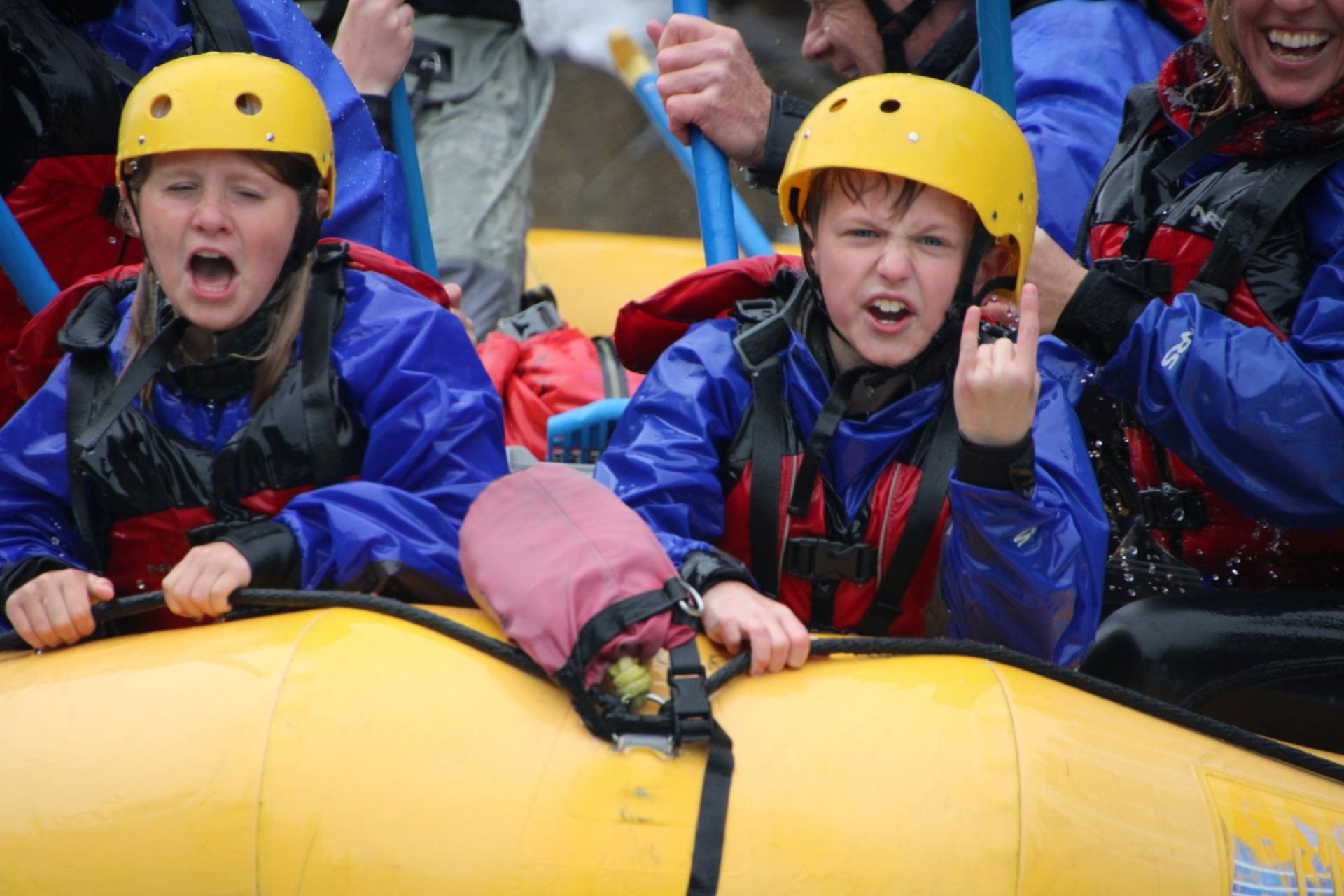 Children in helmets and lifejackets ride a yellow raft through splashing water.