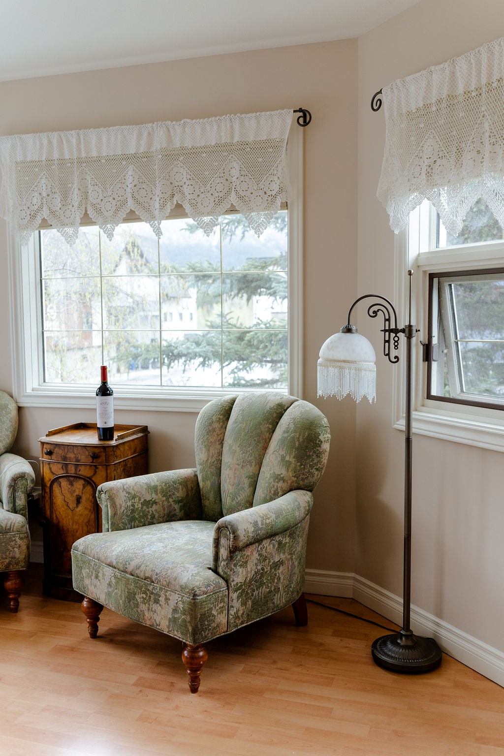 Patterned lace curtains over bay windows, green upholstered armchair, wooden side table, floor lamp.