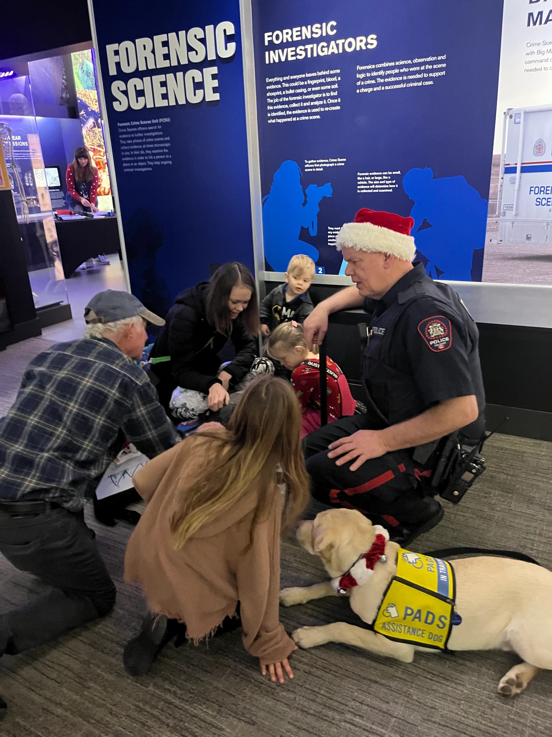 Police officer in Santa hat kneels beside visitors and a PADS assistance dog.