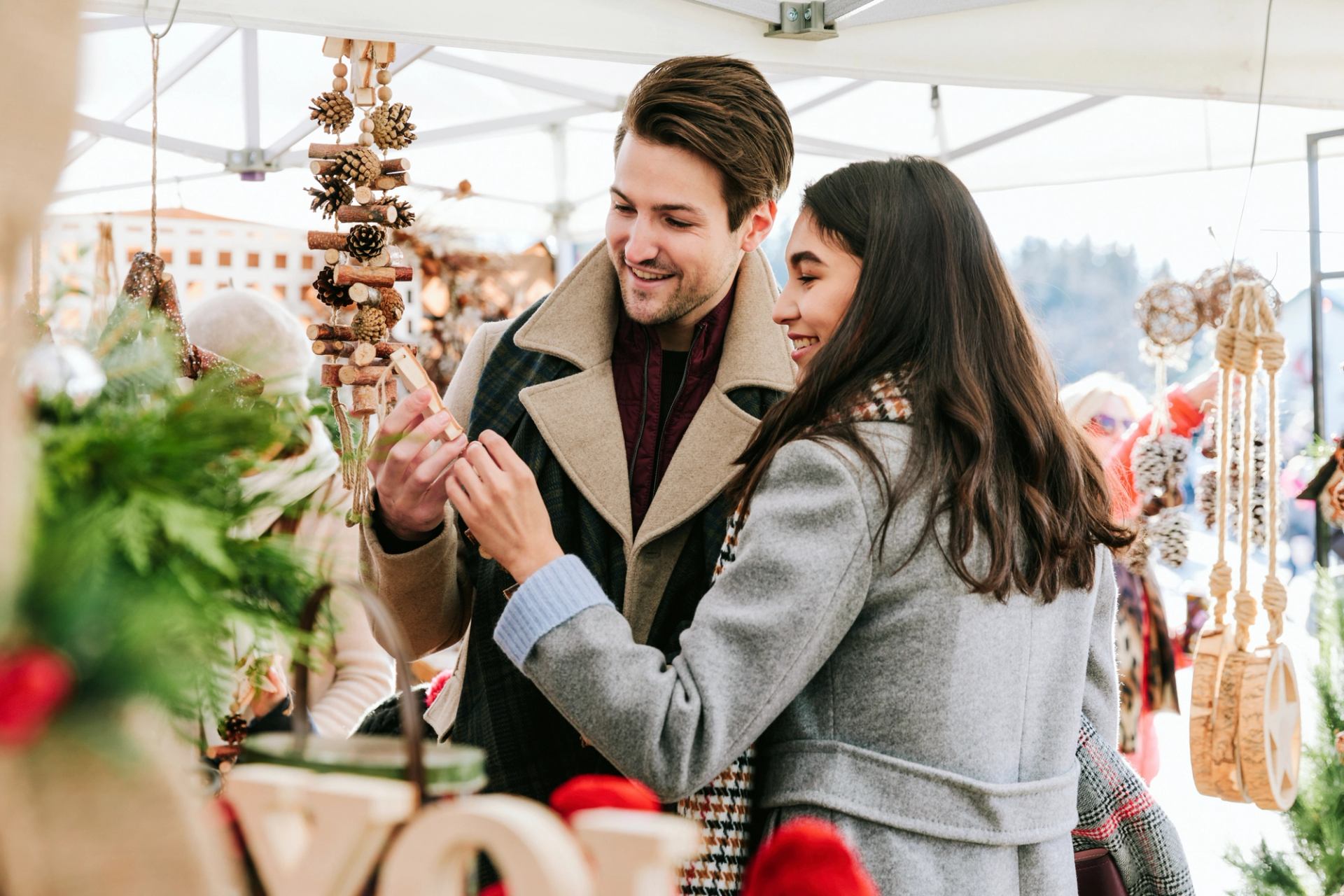 Shoppers examine a small ornament at a market stall with hanging pine cone decorations.