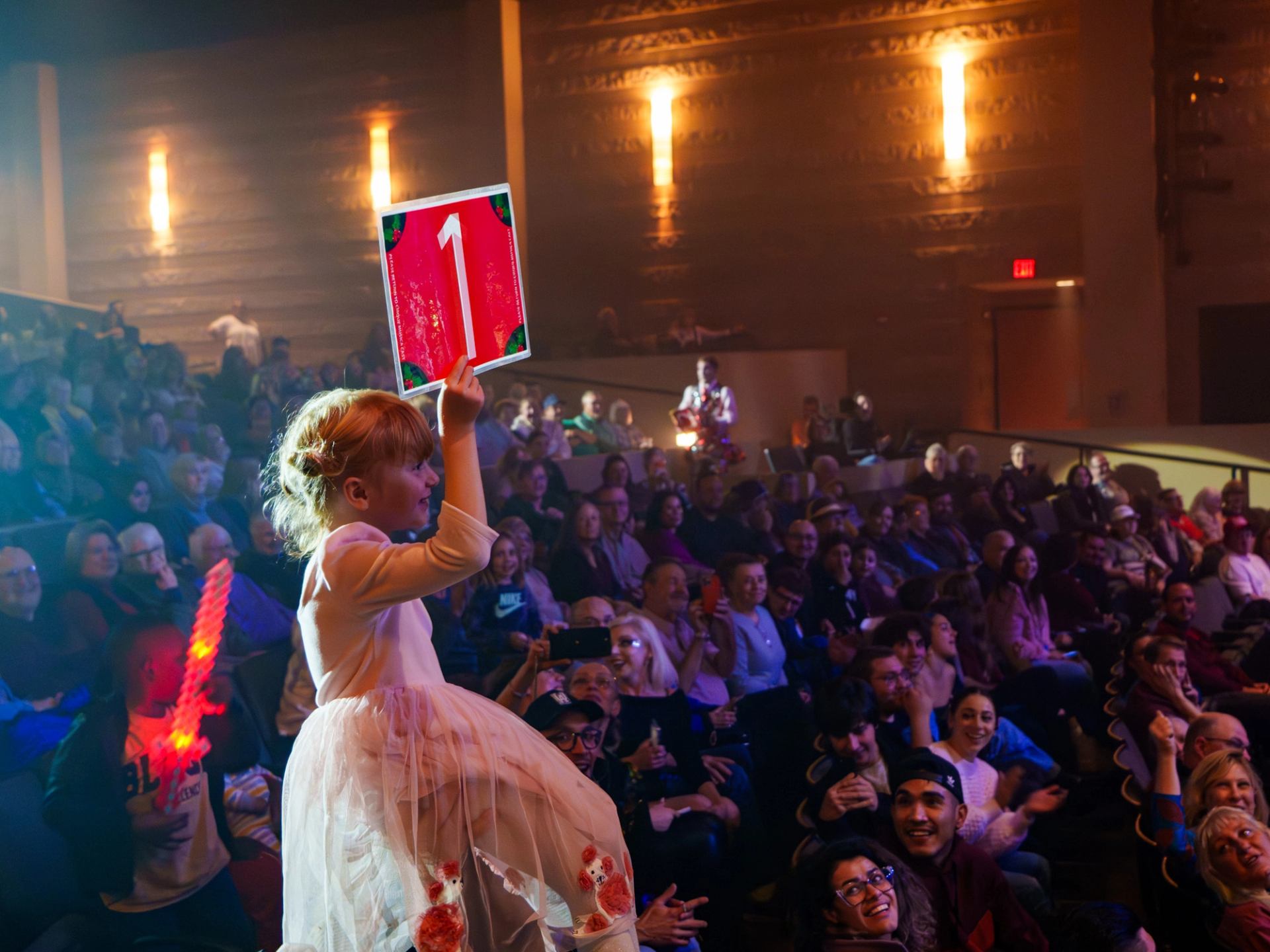 Child holds a red number one sign before a seated theatre audience.