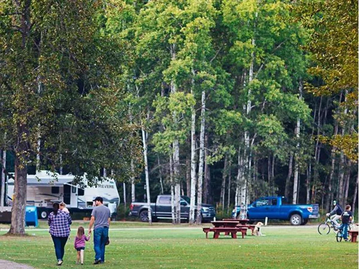 Campground with people walking and cycling near RVs, trucks, picnic tables and tall trees.
