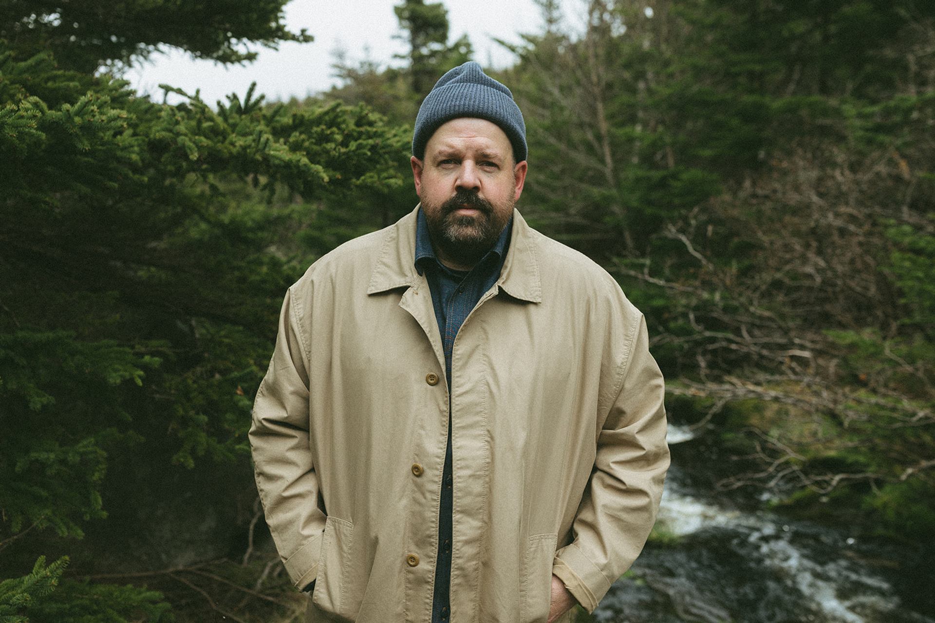 Person in beige coat and knit toque stands in front of forest stream.