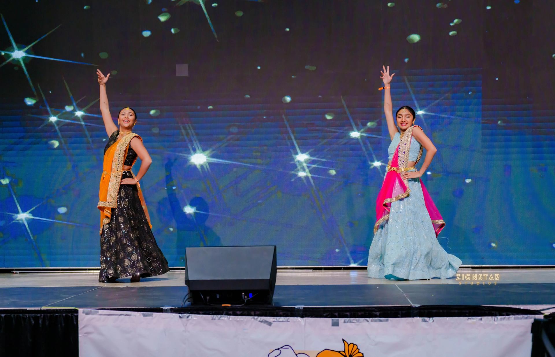 Dancers in colourful traditional attire pose onstage before a sparkling blue backdrop.