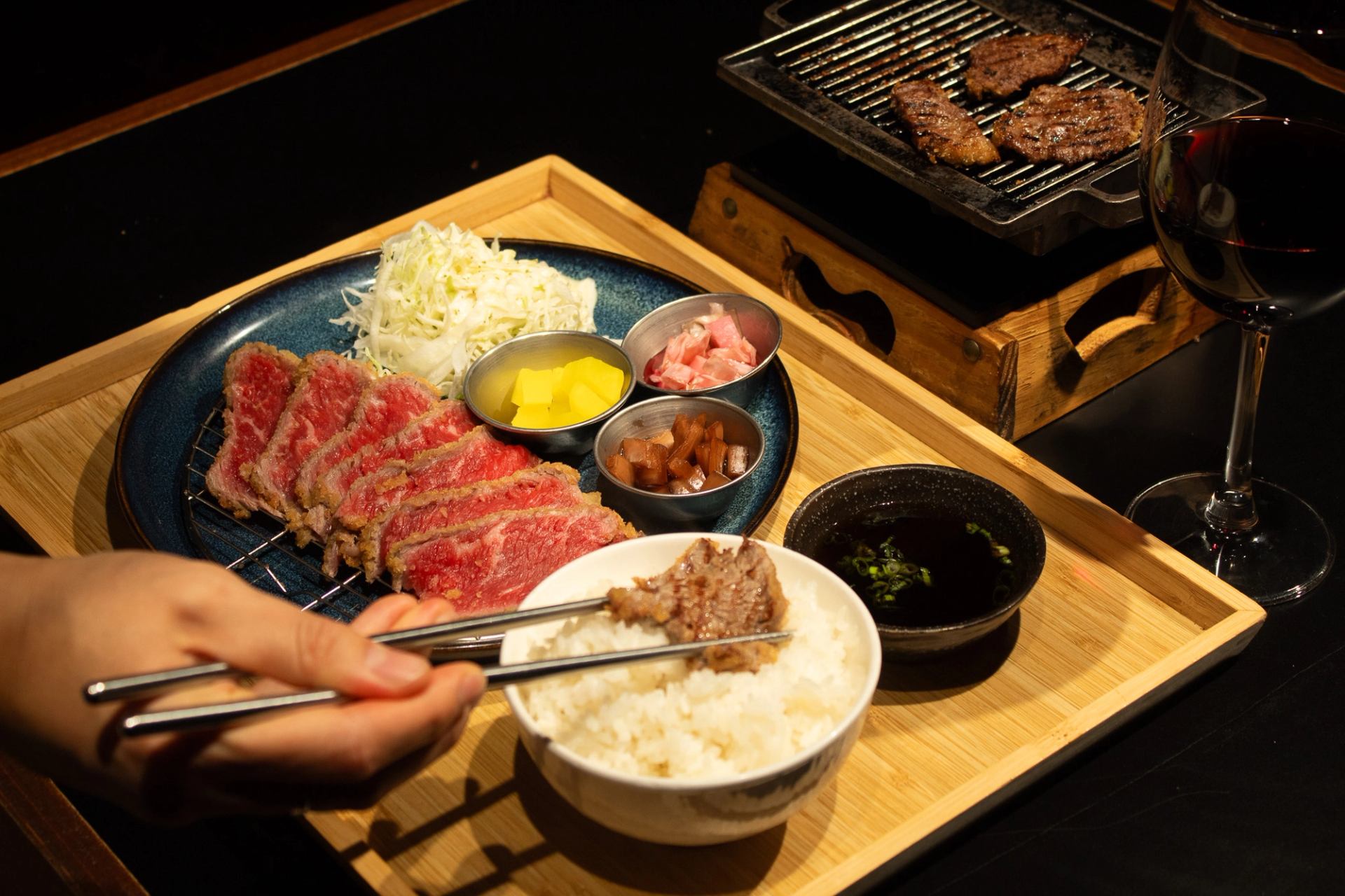 Japanese meal tray with sliced beef, rice, side dishes, tabletop grill and wine.