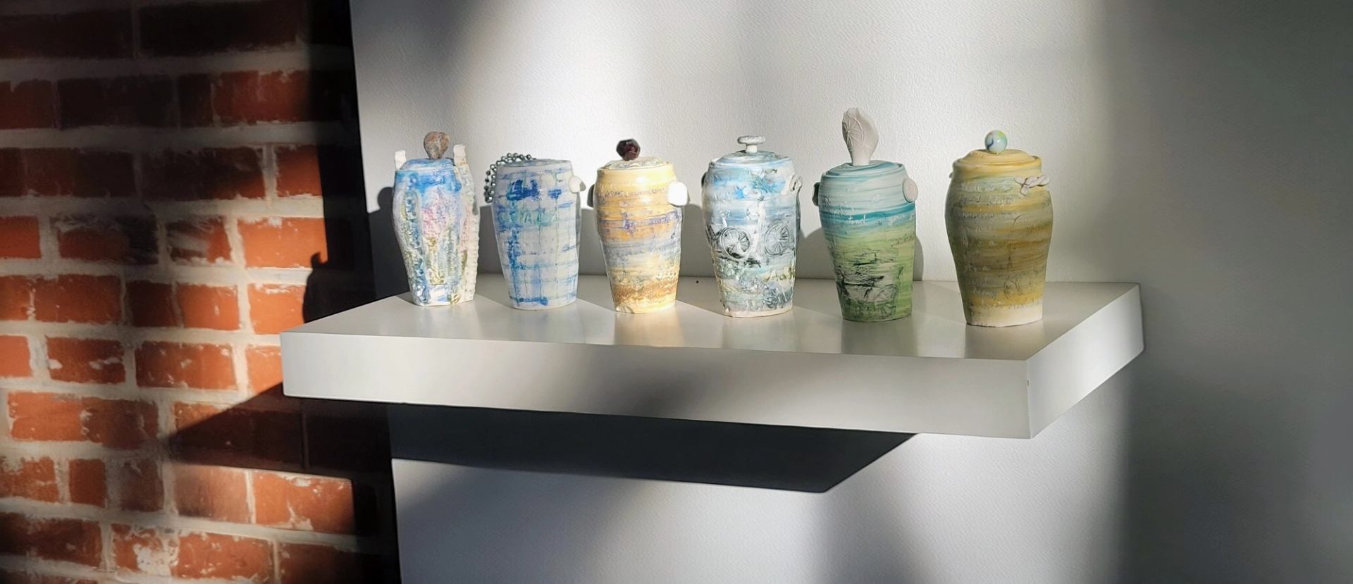 Six unique, colorful ceramic vessels displayed on a white shelf.