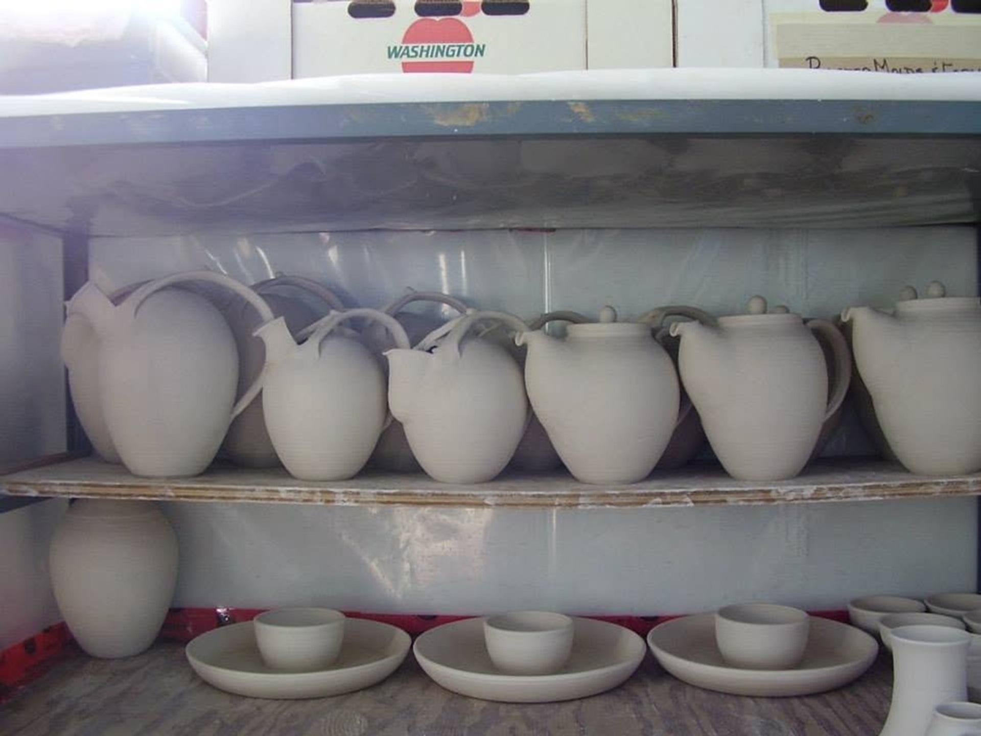Unglazed ceramic pitchers, bowls, and plates are neatly stacked on shelving before firing