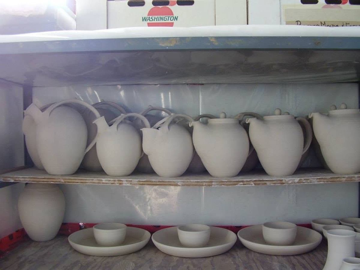 Unglazed ceramic pitchers, bowls, and plates are neatly stacked on shelving before firing