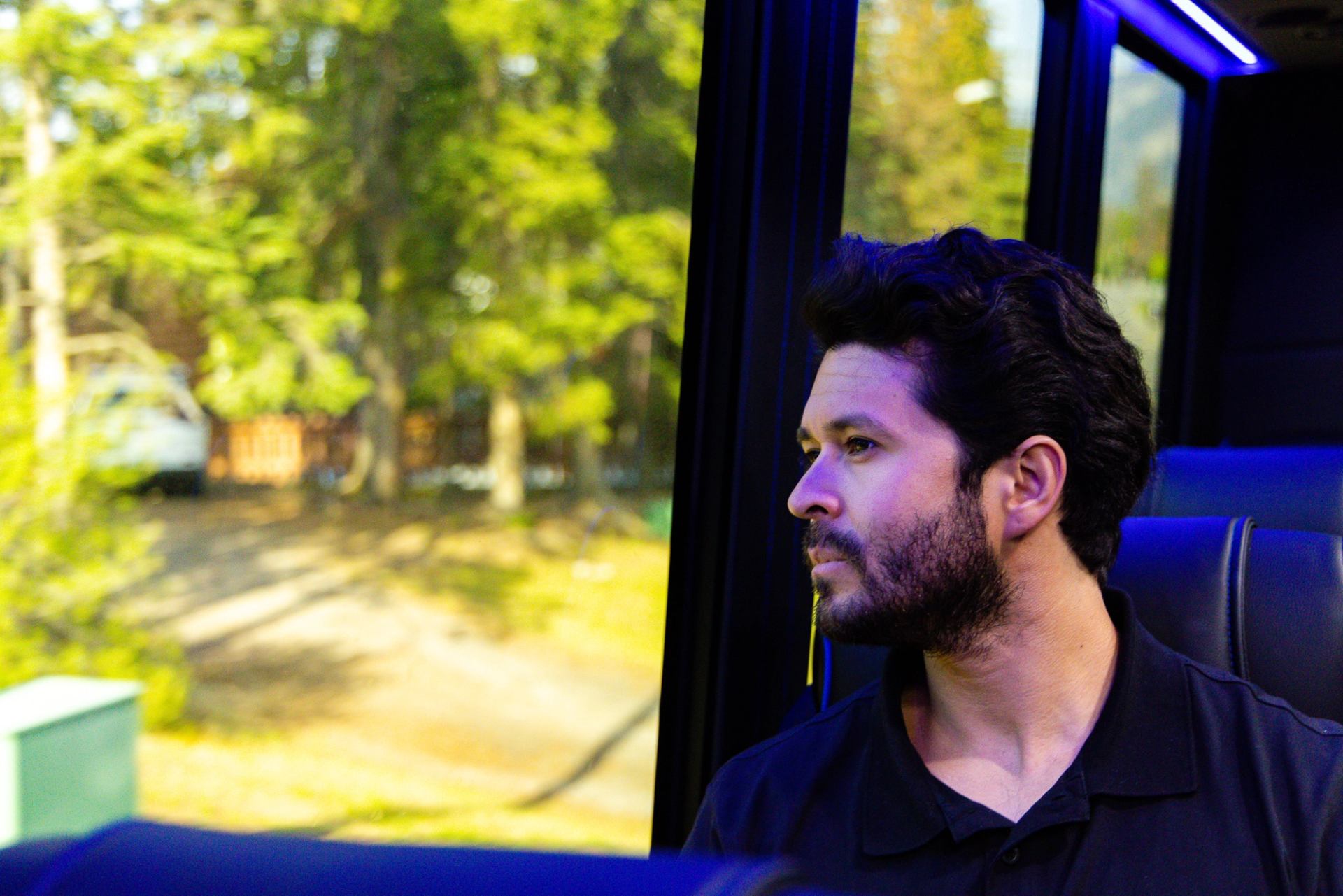 Man sits on a bus, looking out the window at trees and parked vehicles.