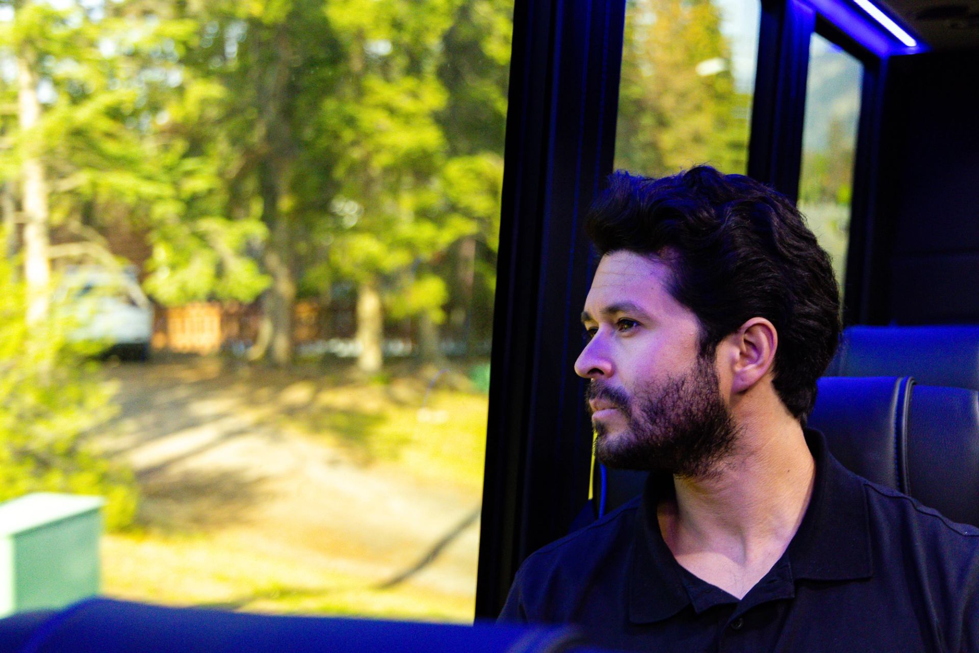 Man sits on a bus, looking out the window at trees and parked vehicles.