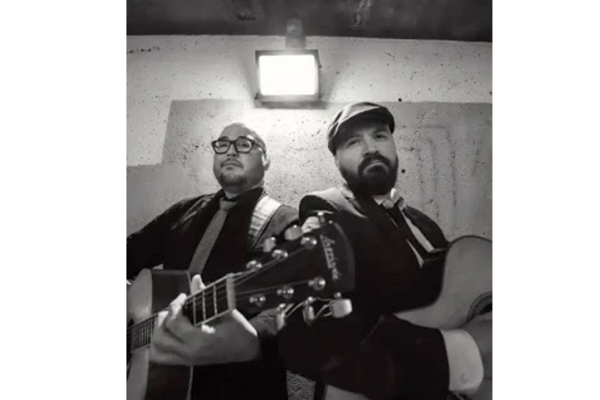 Guitarists perform beneath a wall-mounted light in a black-and-white image.