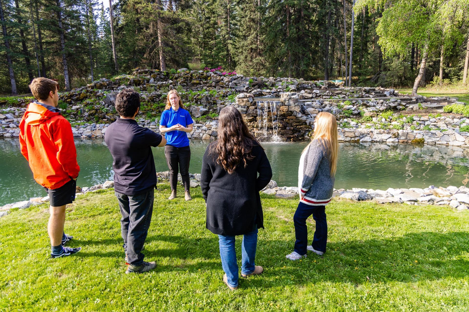 Guide speaks to small group beside pond with rock waterfall and forest backdrop.