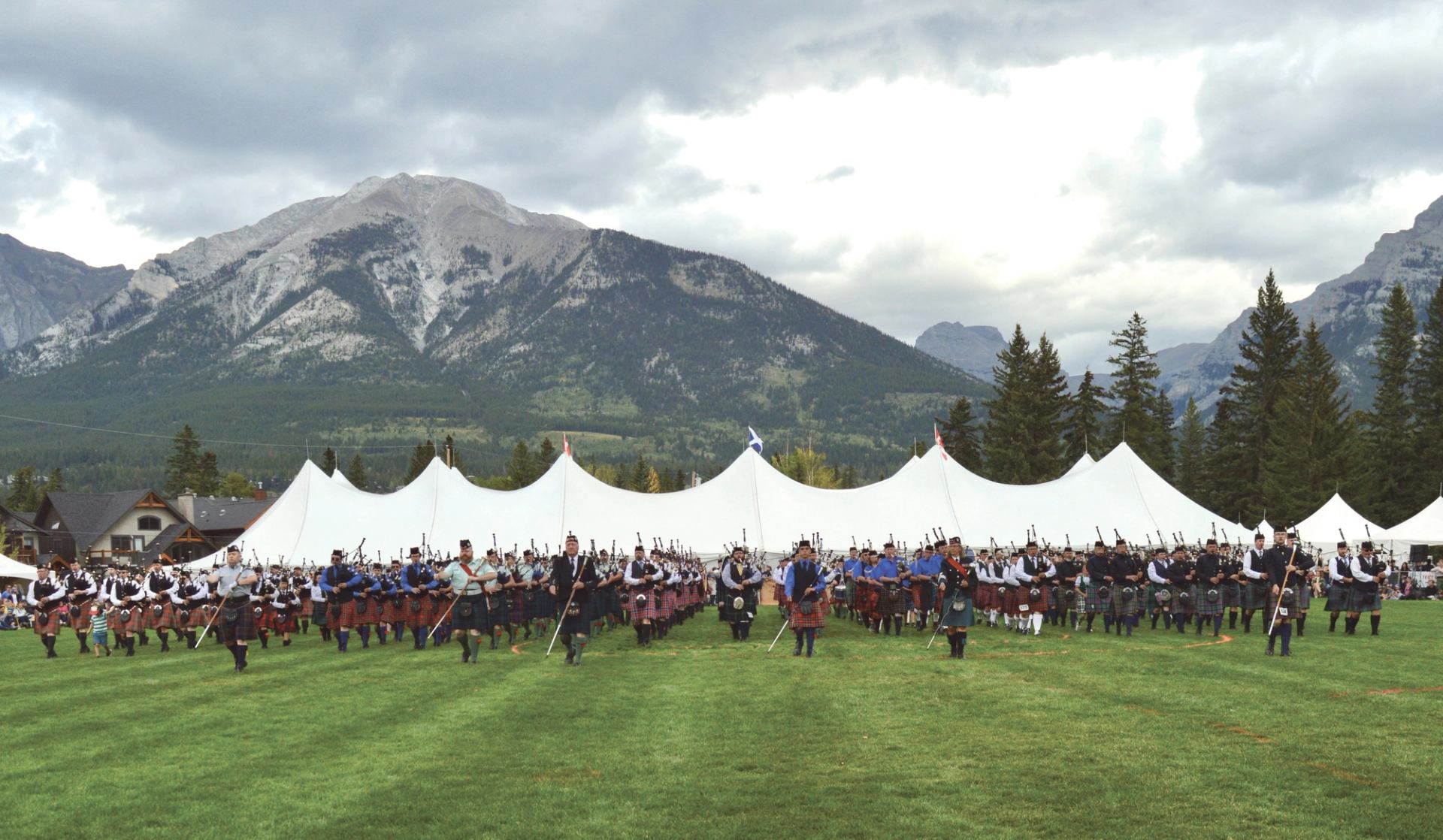 Pipe band marching in front of white tents with mountains and cloudy sky behind.