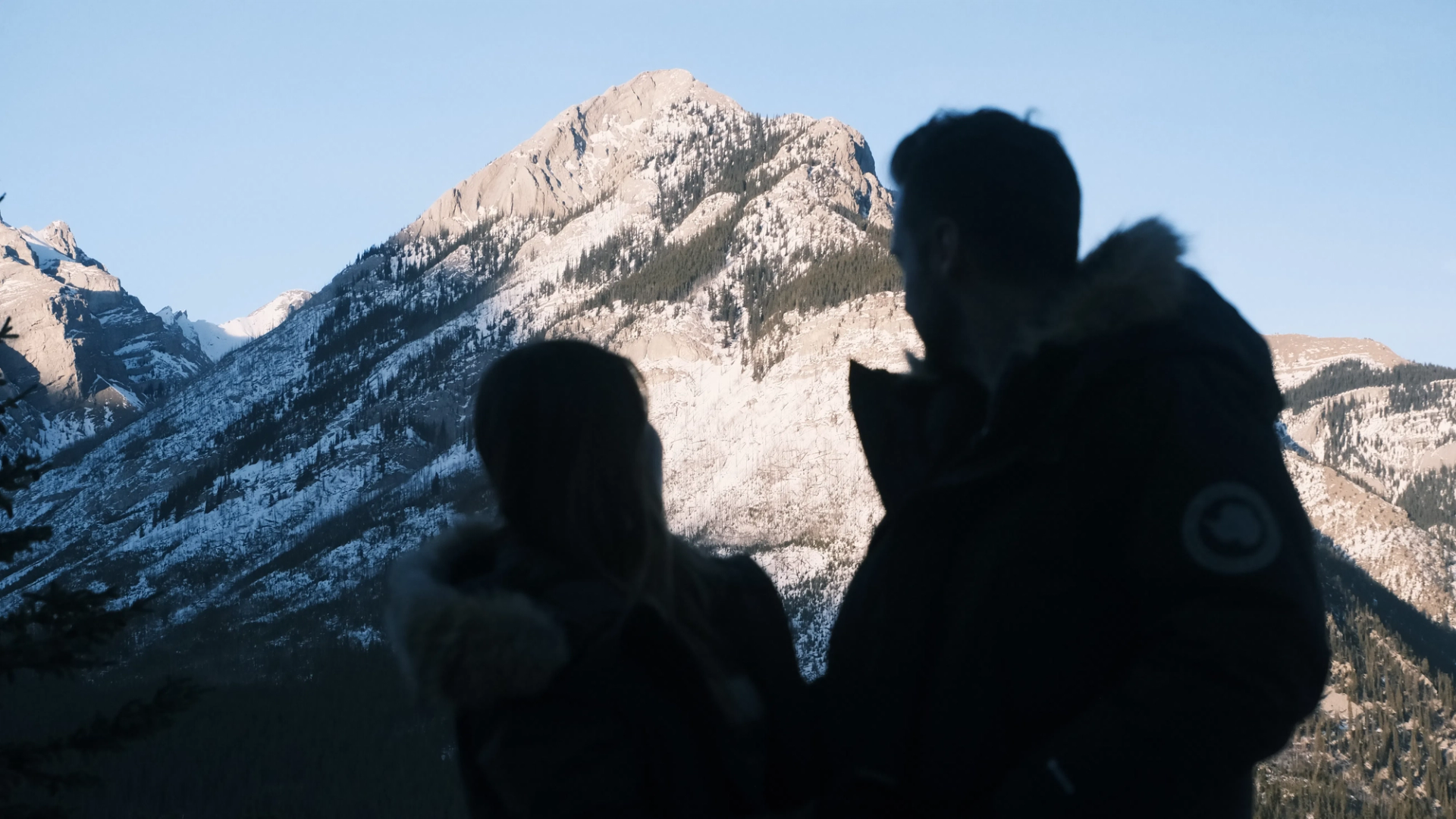 Silhouetted hikers in winter jackets stand before a snow-dusted mountain landscape.