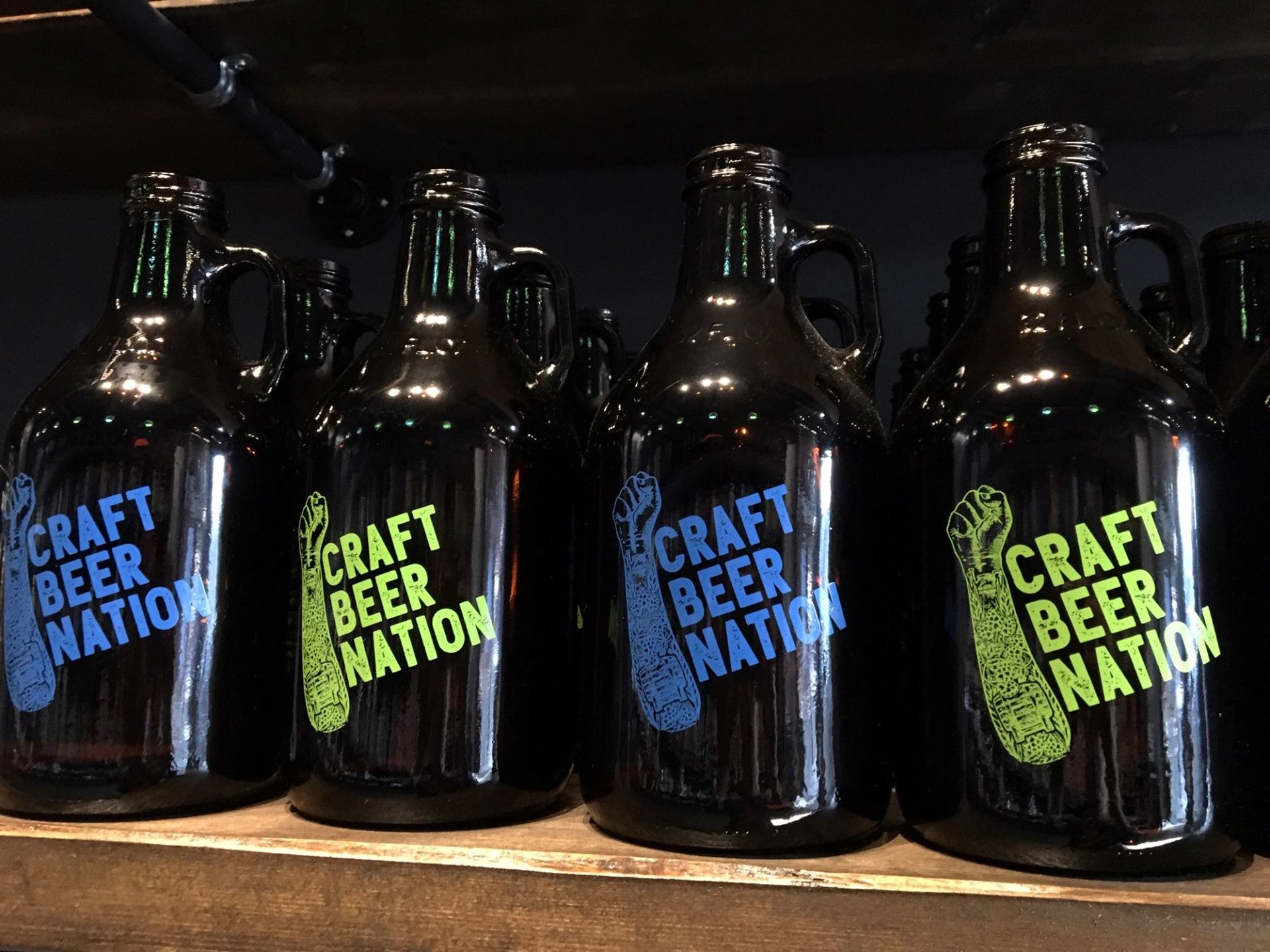 Row of Craft Beer Nation growlers in blue and green on a wooden shelf.