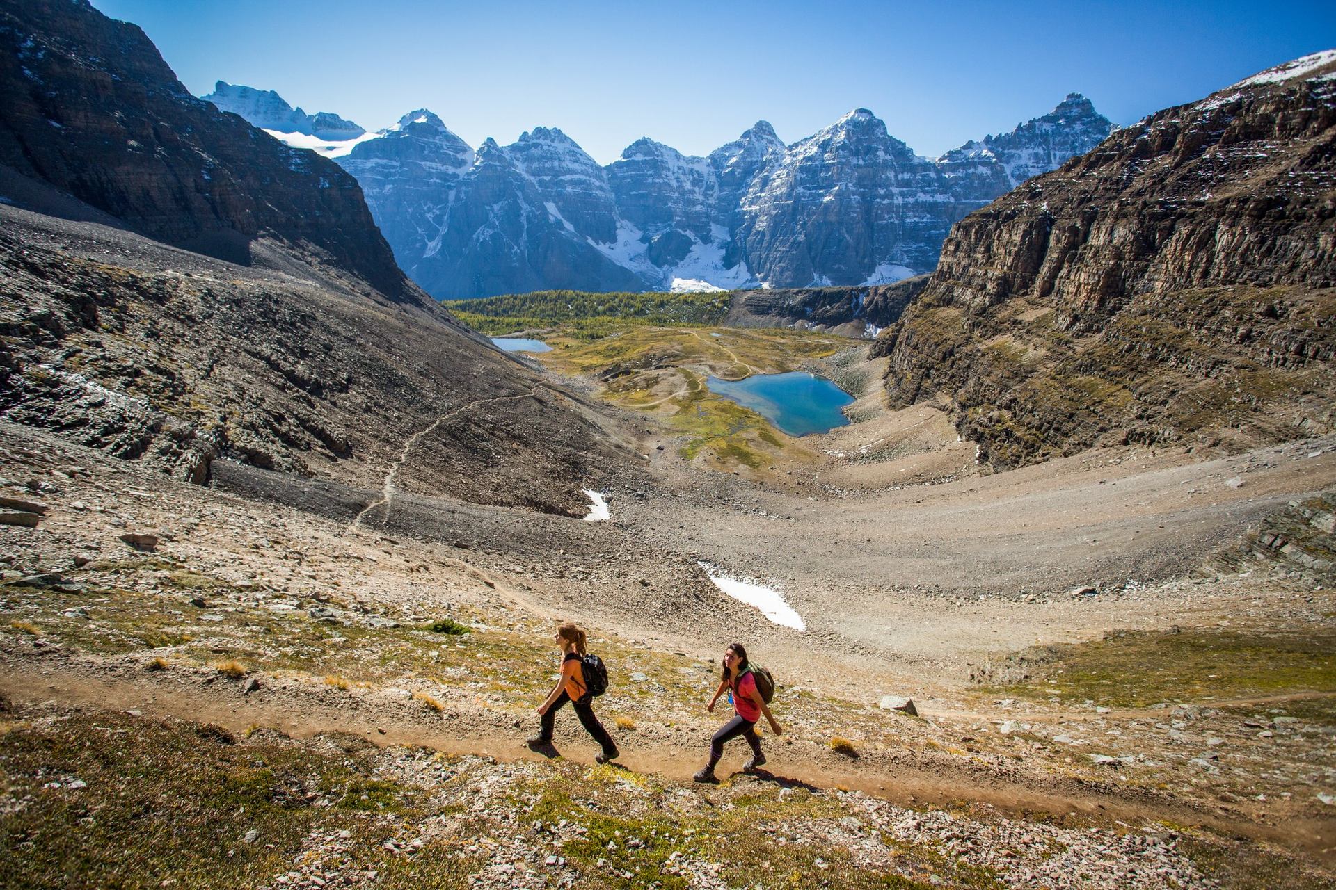 Two hikers on a mountain trail in a vast valley with blue lakes and snowy peaks.