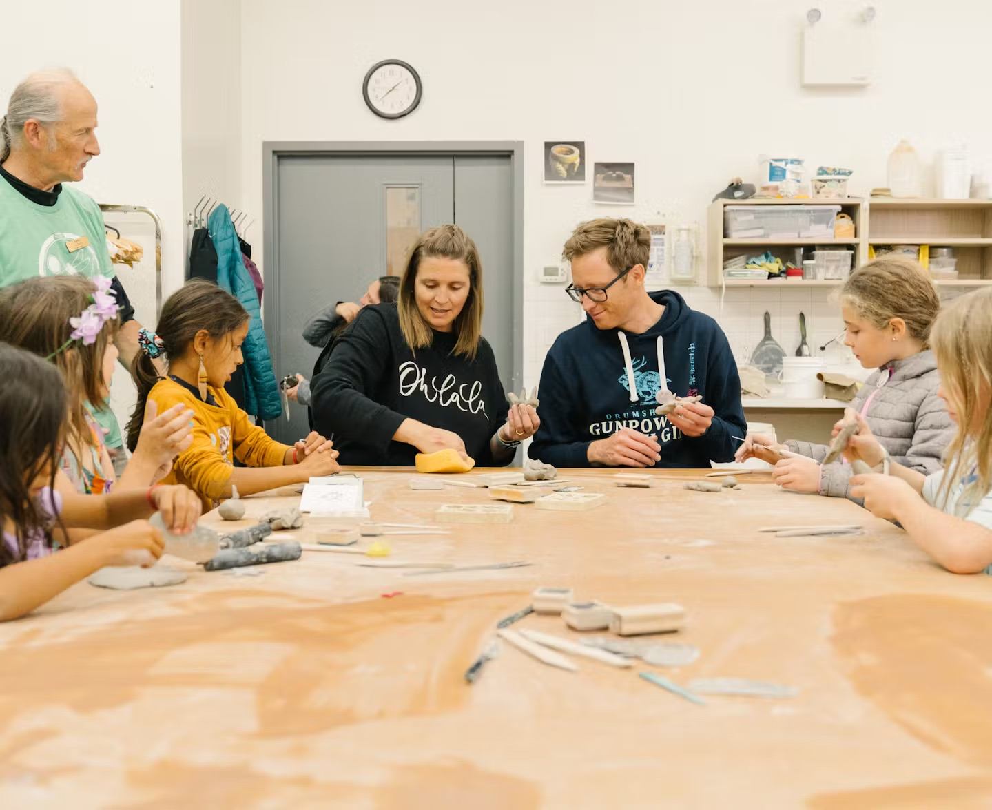 Children and adults gather around a table for a hands-on clay workshop in a studio setting.