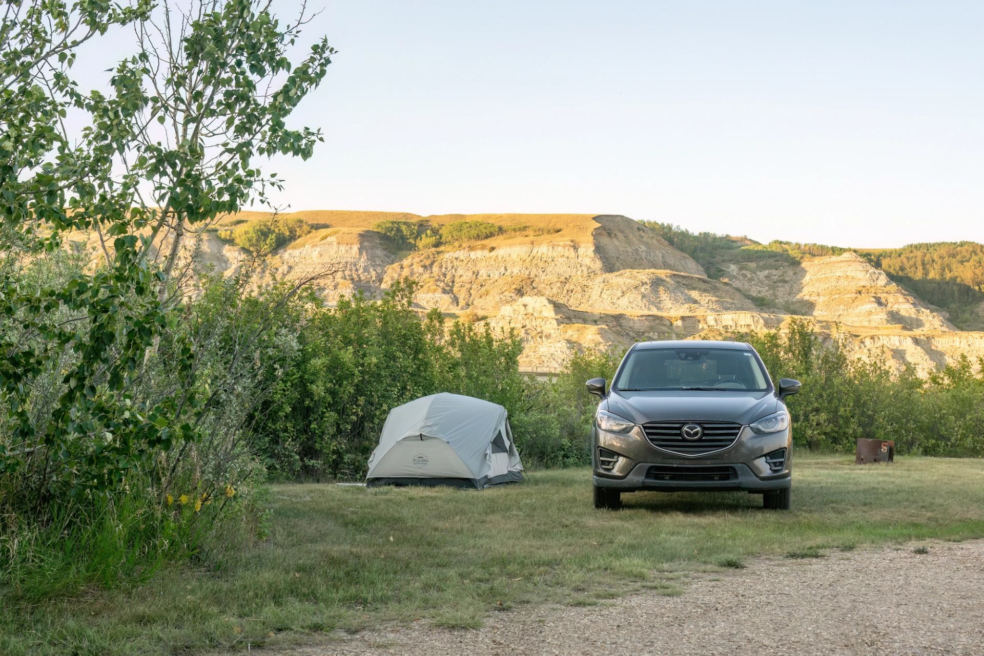 Campsite with grey tent and parked car in grassy area below rocky hills.