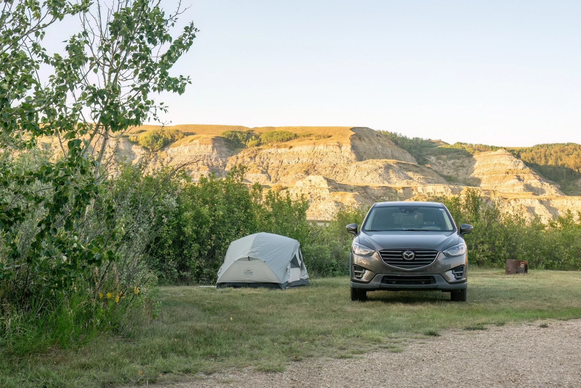 Campsite with grey tent and parked car in grassy area below rocky hills.
